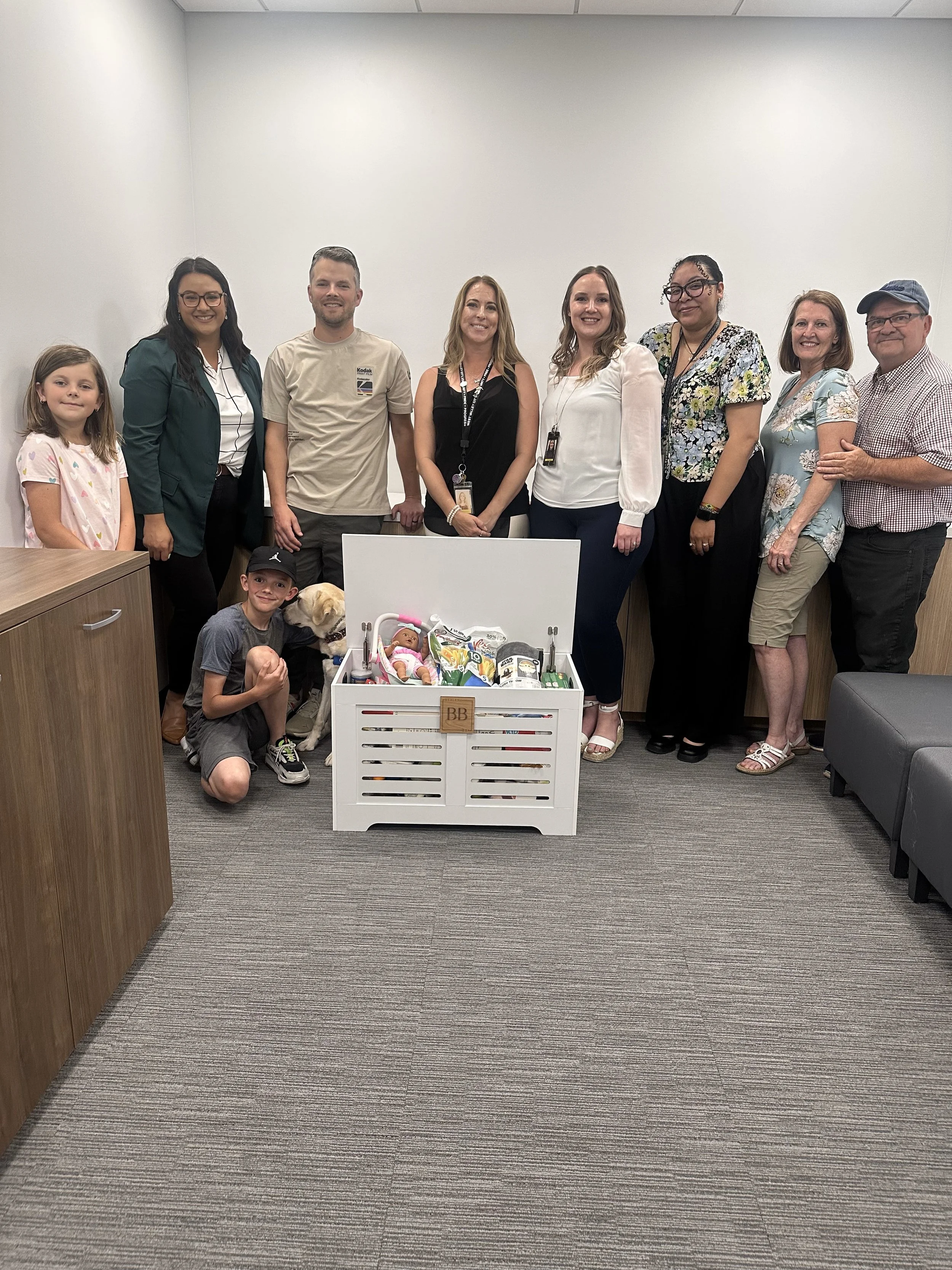 Group of nine adults and two children posing with a small crate filled with baby supplies and toys in a plain office room.