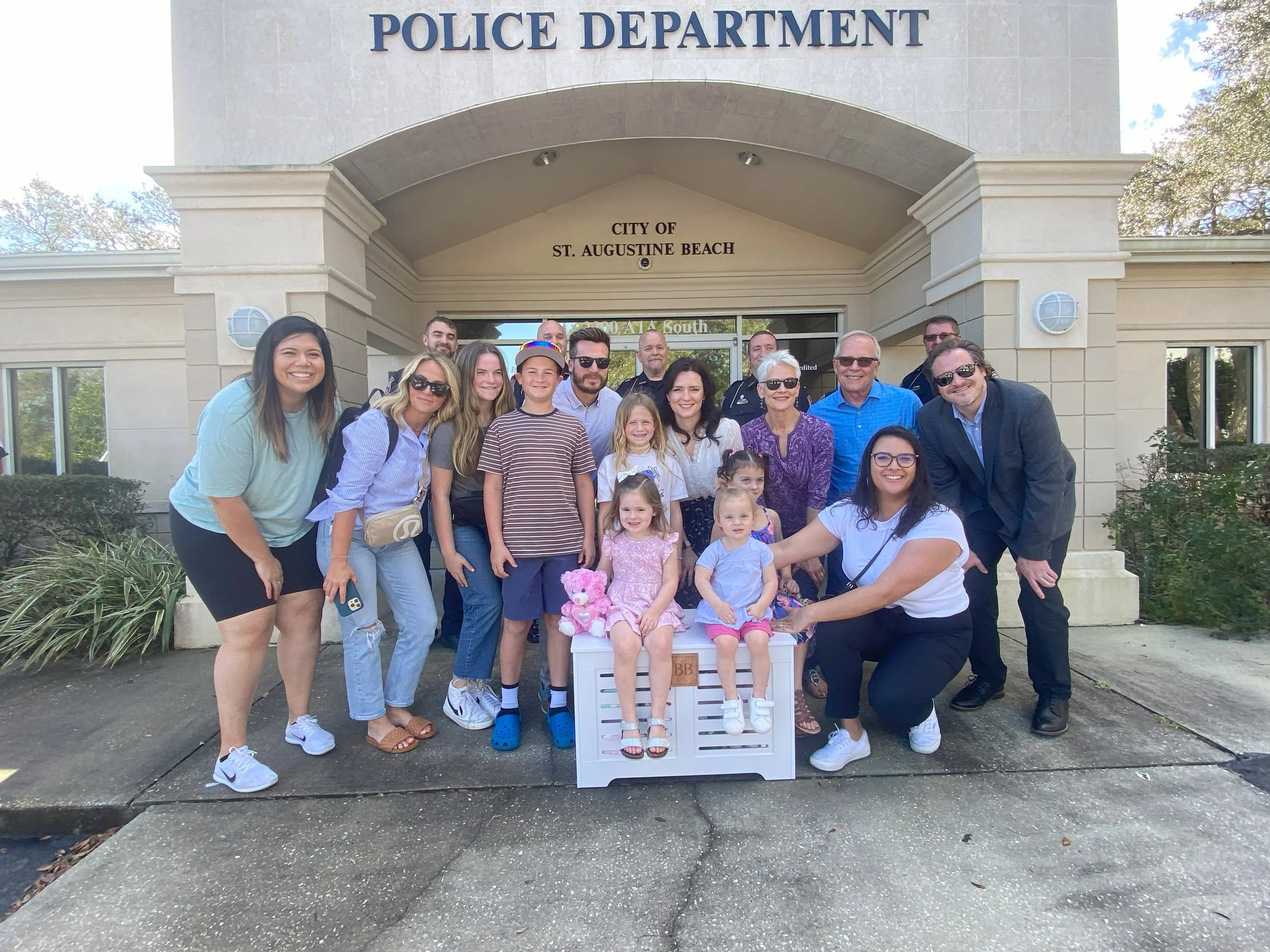 A group of people, including children and adults, posing in front of the police department building in St. Augustine Beach, Florida. They are smiling and gathered around a small white dresser with children sitting on it, some holding stuffed animals.