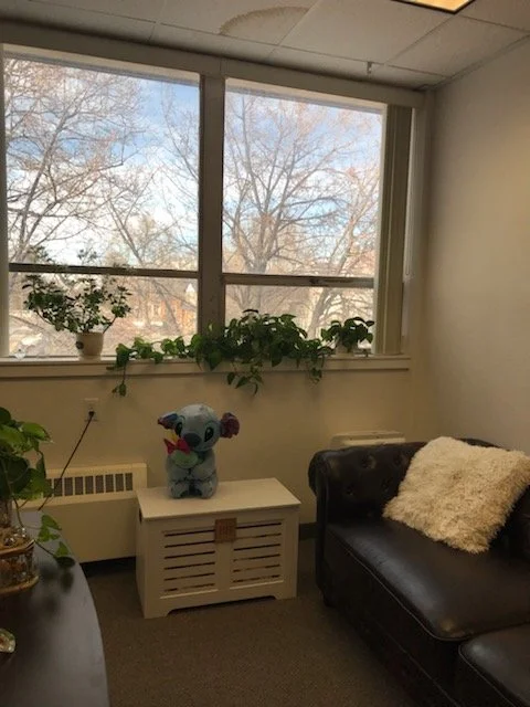 An office room with large window, green plants on windowsill, plush toy of Stitch from Lilo & Stitch on a white table, black sofa with white fur pillow.