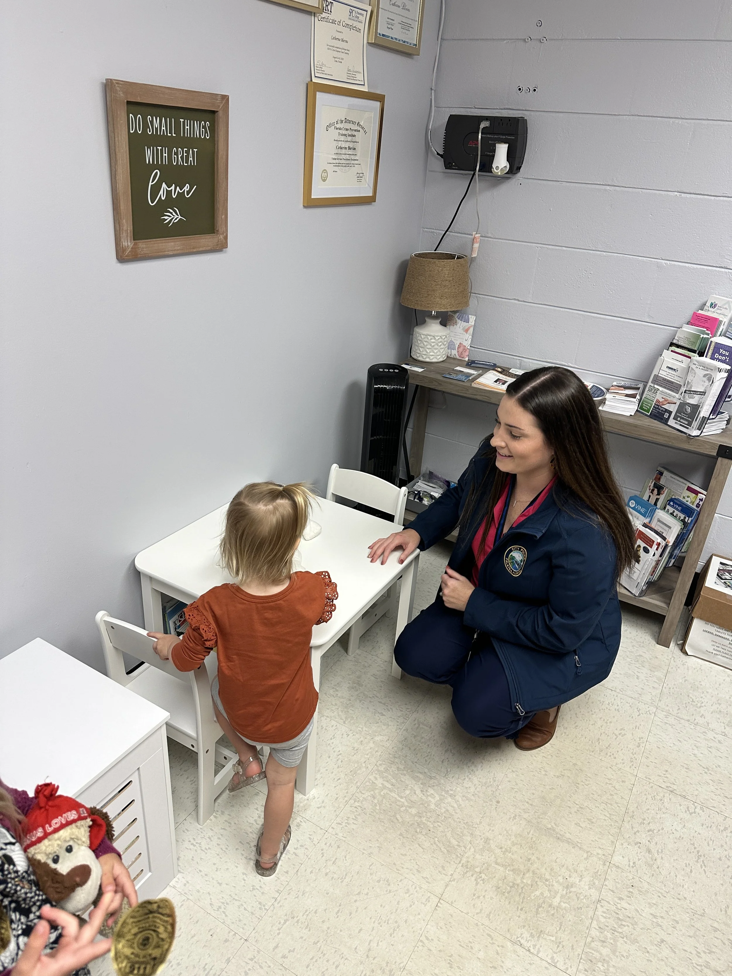 A woman in a dark blue jacket kneeling and talking to a young girl with blonde hair, wearing an orange top and shorts, in a small room with framed certificates on the wall.
