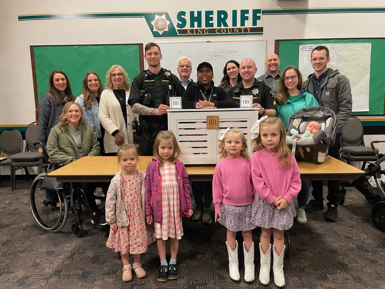 Group photo of sheriff officers, children, and adults in a room with a sheriff's office sign, some children are wearing boots, and a baby is in a car seat, with a white chest in front of officers.