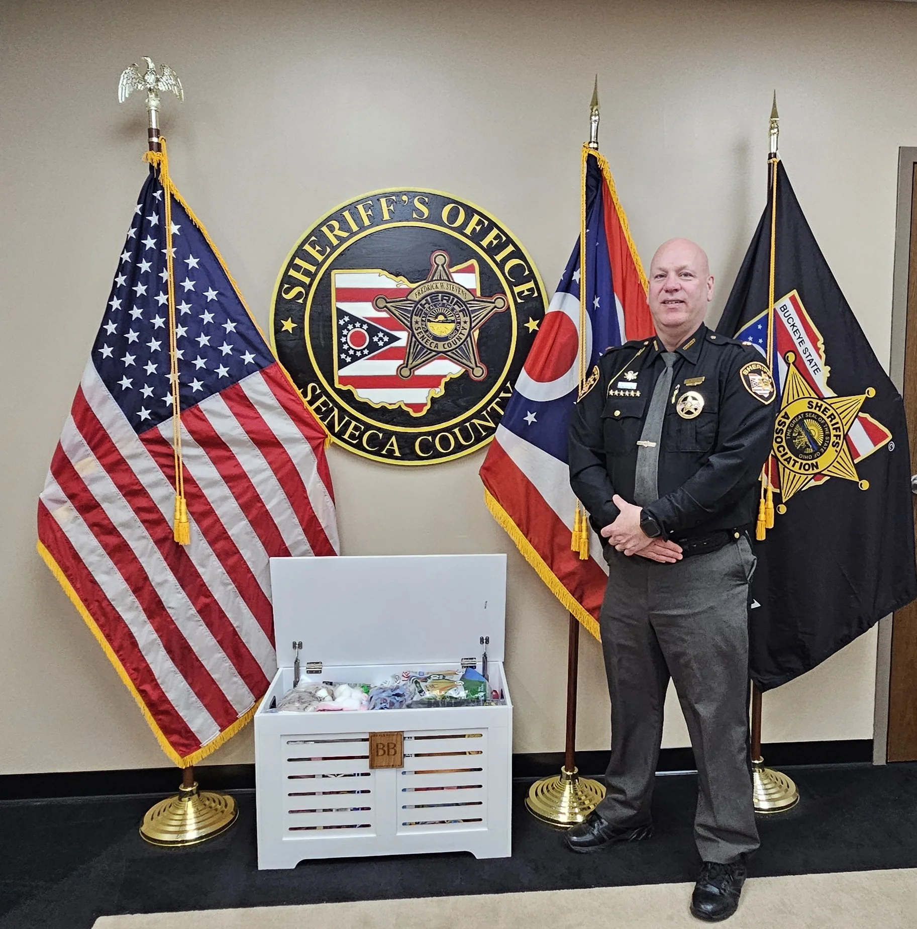 A sheriff's officer standing in front of four flags, including the American flag and a sheriff's office emblem, next to a white crate filled with small items.