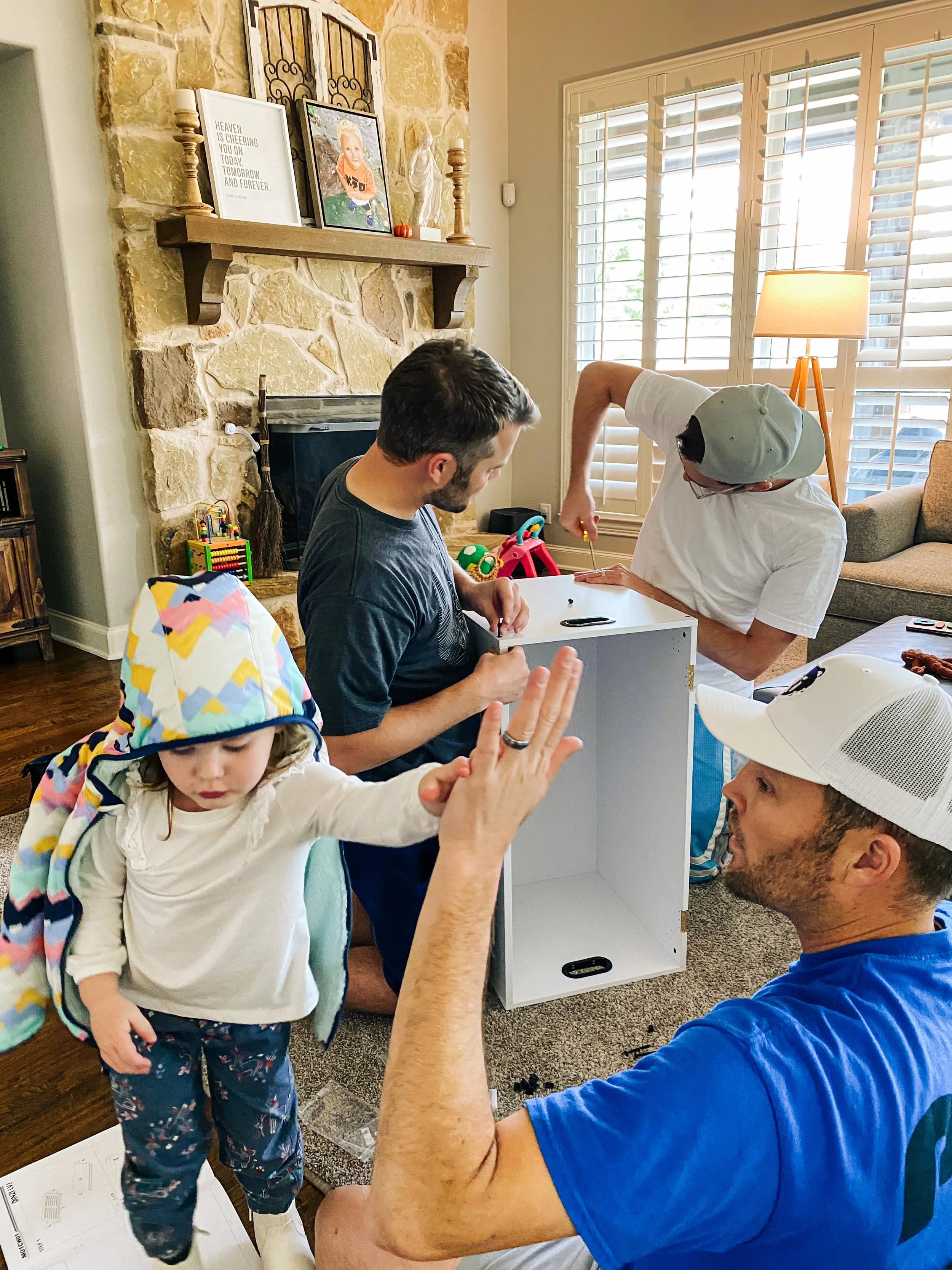 A family of four assembling furniture in their living room; a young girl wearing a colorful hoodie is reaching out with her hand to high-five an adult man, while another adult man in a white shirt and gray cap is working on the furniture, and a woman