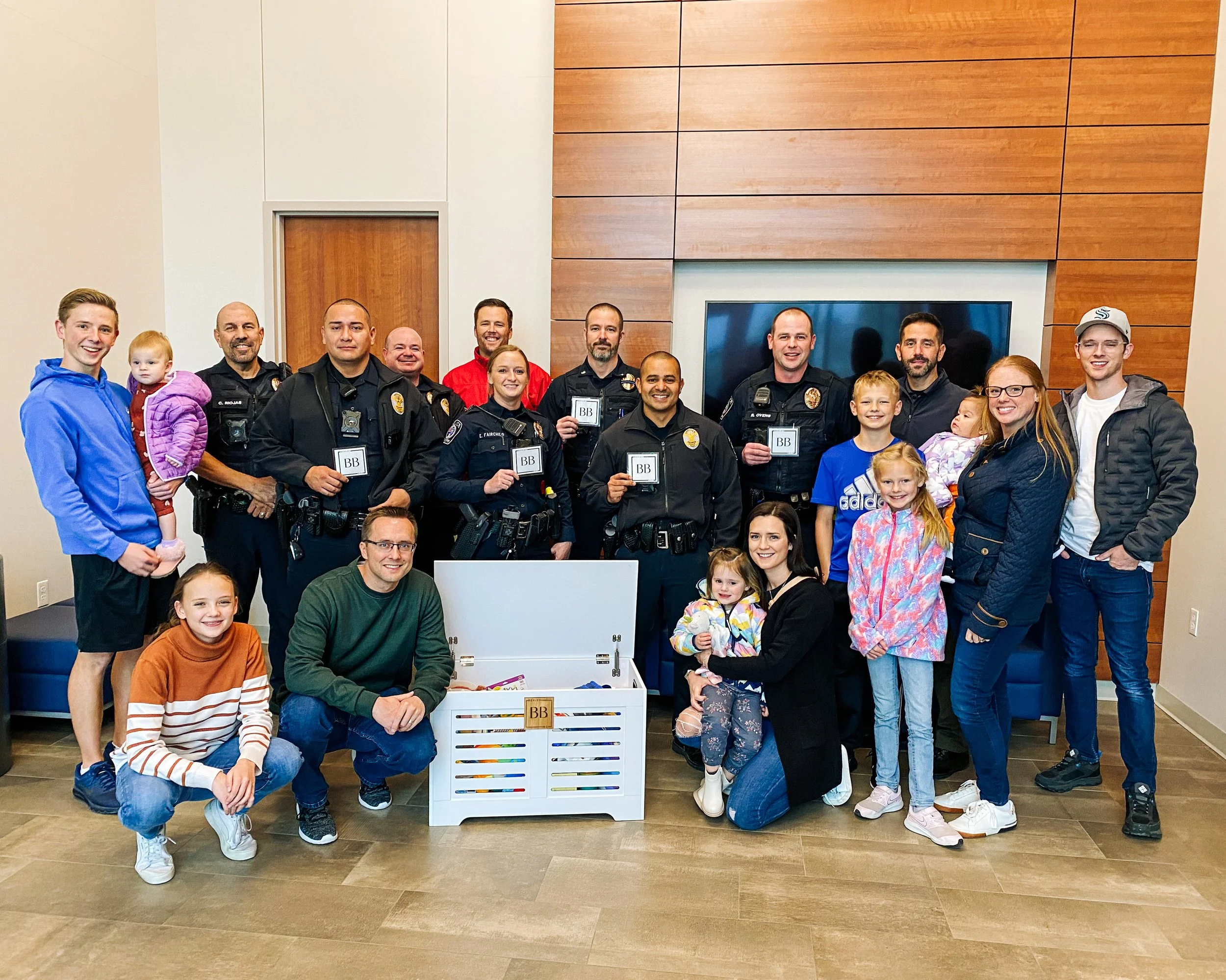 Group photo of police officers and families indoors, children and adults smiling, with a large television screen and wooden panel wall in the background, and a white crate with brightly colored pencils in the foreground.