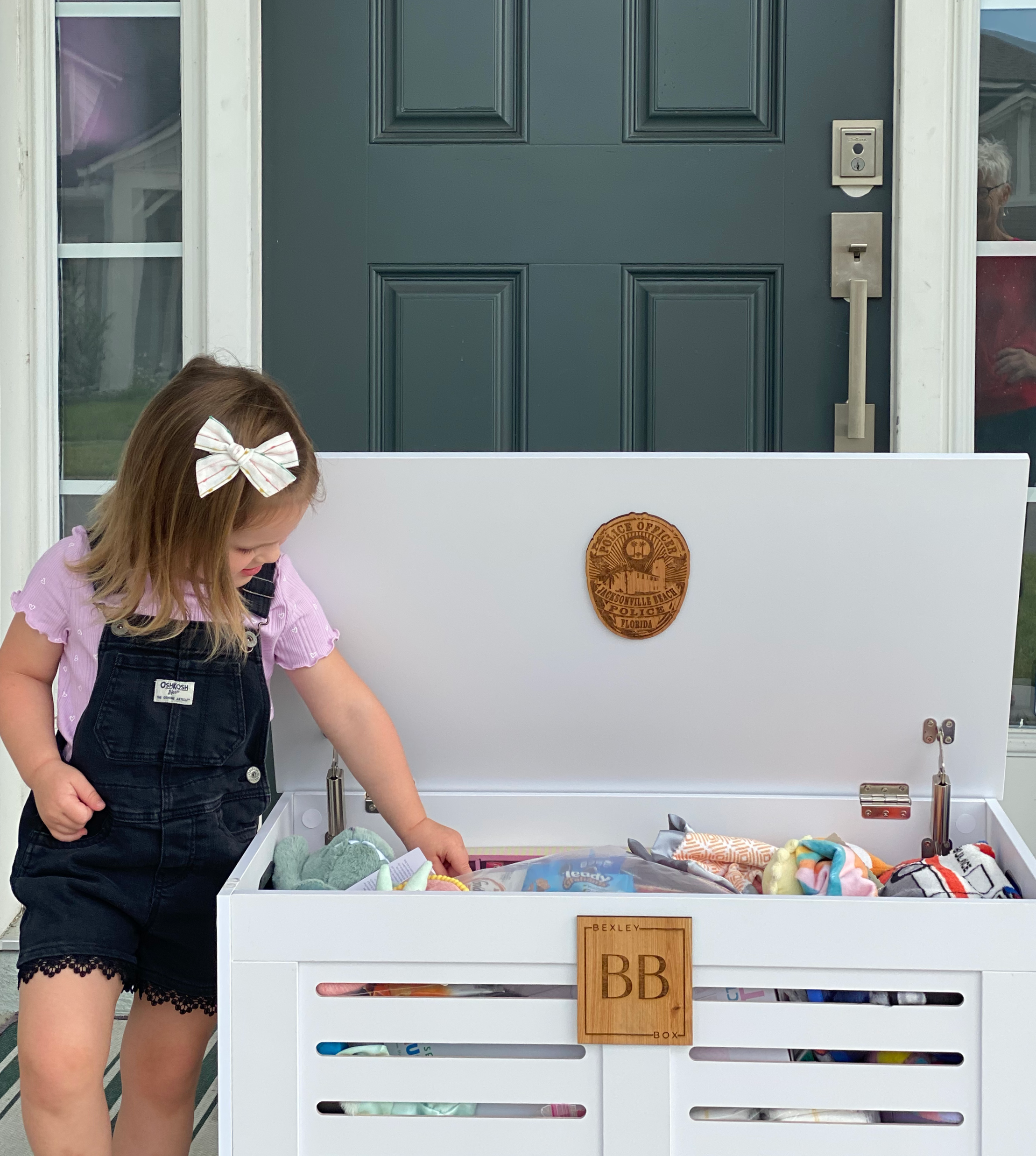 A young girl with a white bow in her hair, wearing a pink shirt and black overalls, is reaching into a large white toy trunk filled with toys. The trunk is outside a house with a blue front door and a porch, and a person is visible through a window on the right.