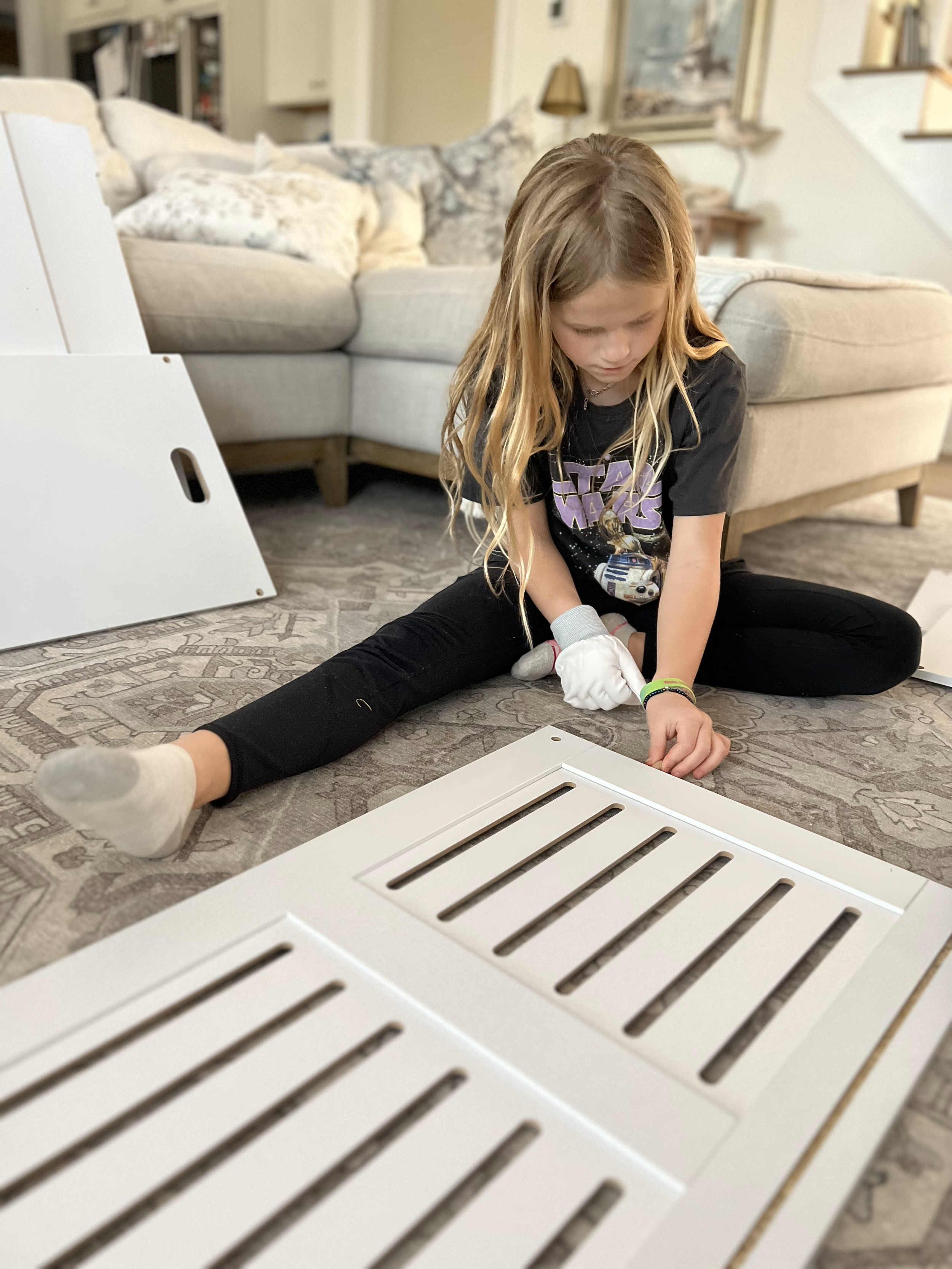 A young girl sitting on the carpeted floor, assembling a white vent cover in a living room.