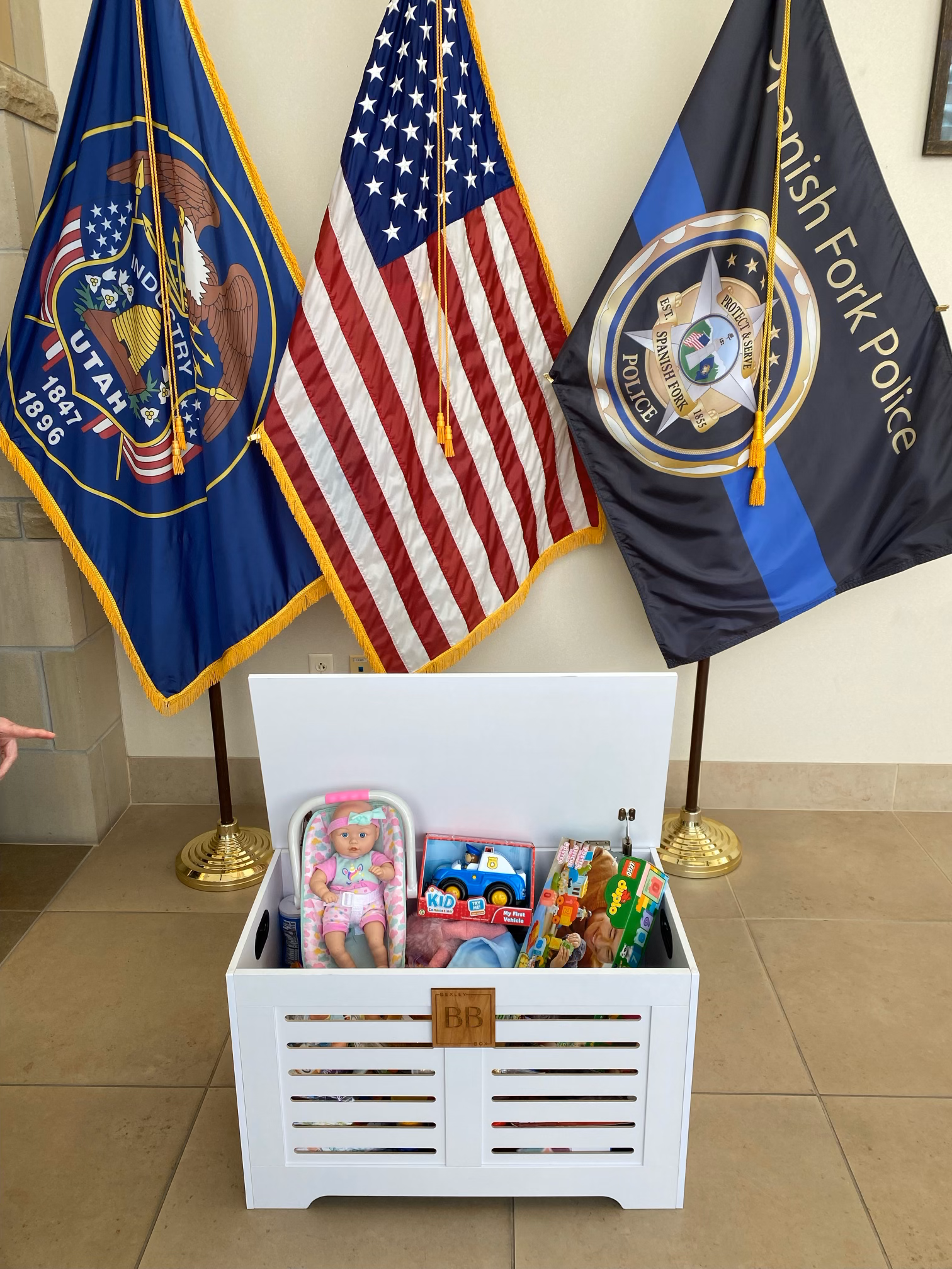 A collection of flags, including the United States flag and two police department flags, positioned behind a white toy chest filled with children's toys and a doll, on a beige tile floor.