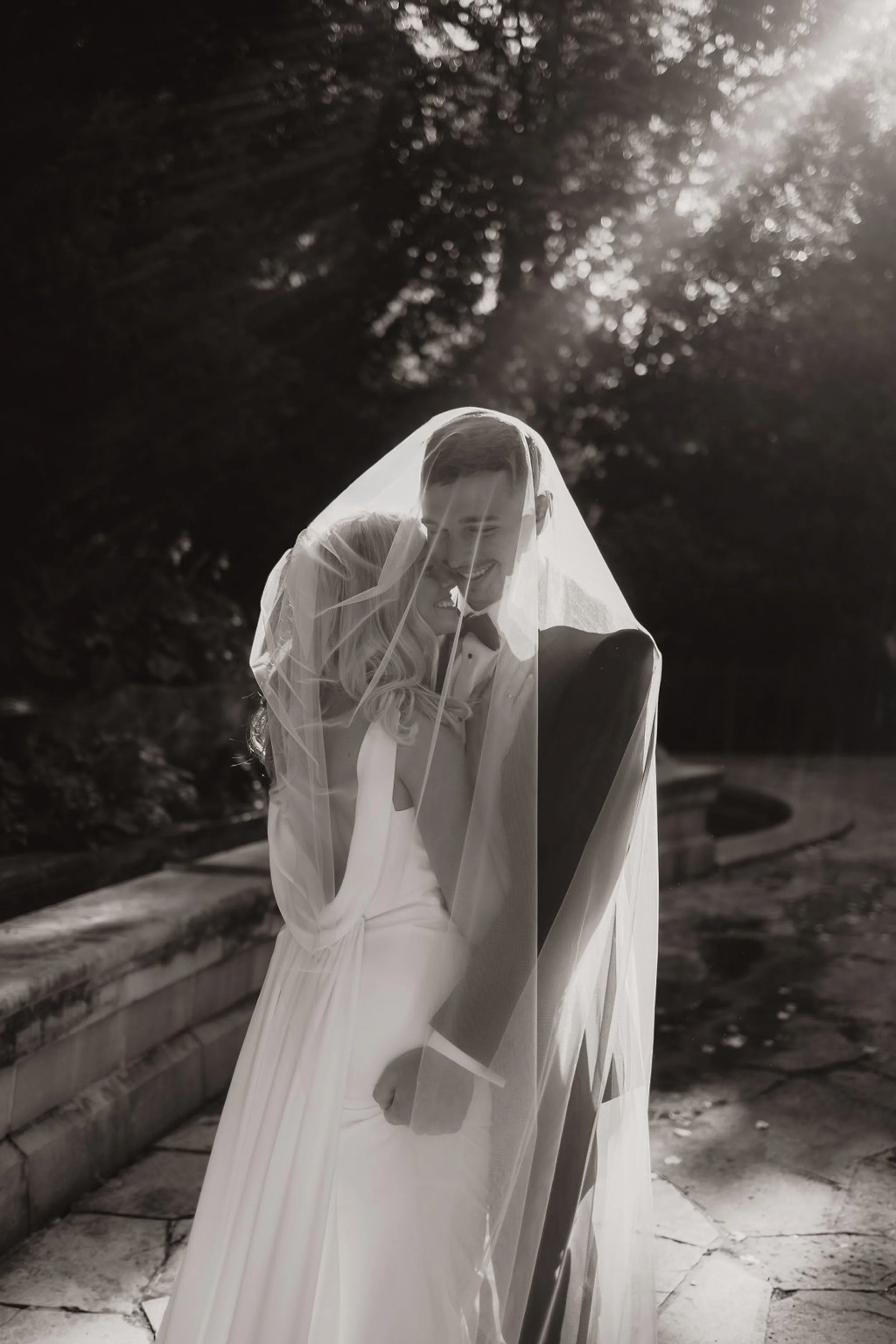 A bride and groom under a veil smiling at each other during their wedding photos outdoors in black and white. Empire Room Omaha Nebraska