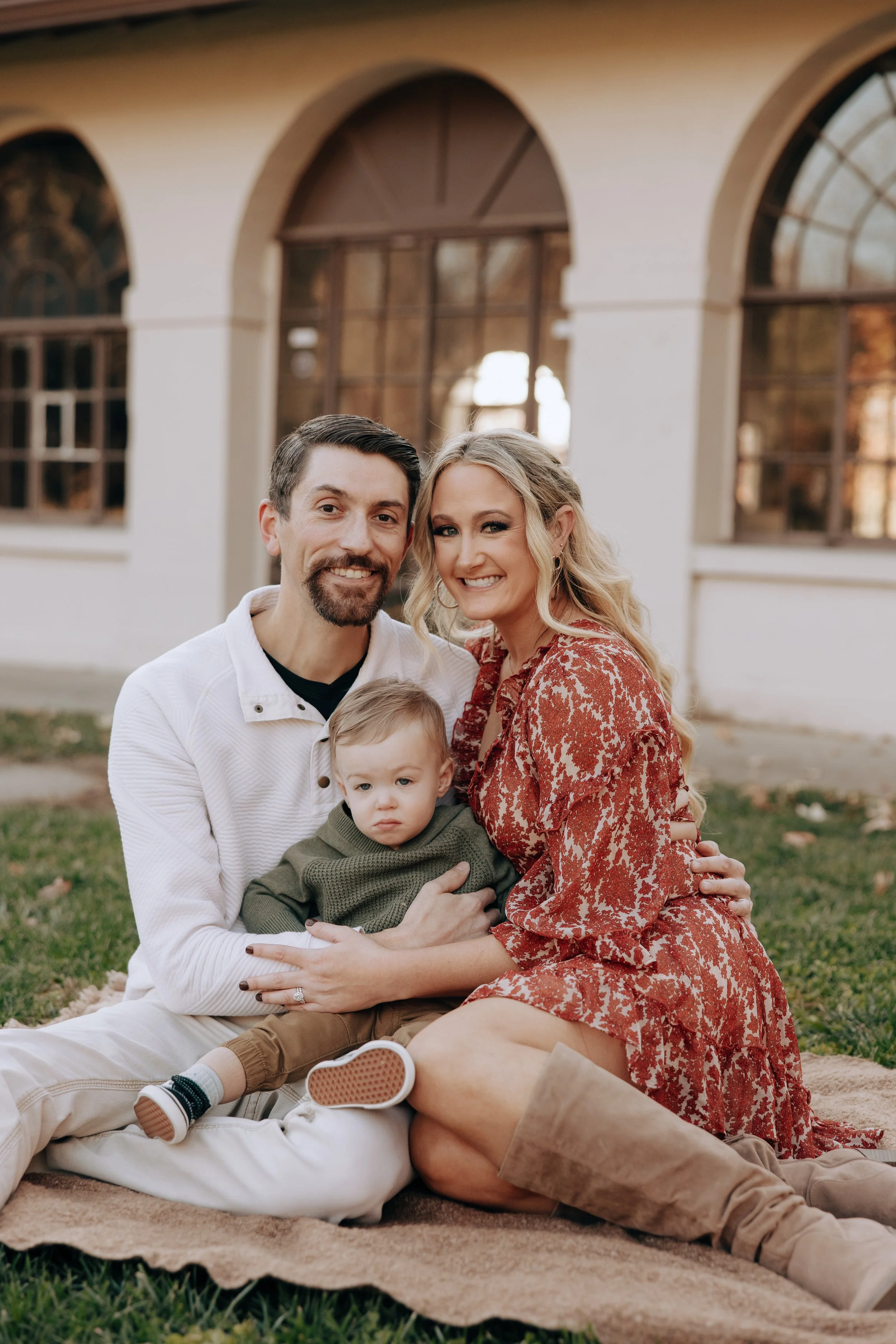 Family Photographer Katelyn Rowan Photography Omaha Nebraska. A family of three sitting on a blanket outside in front of a building with large arched windows, smiling at the camera.