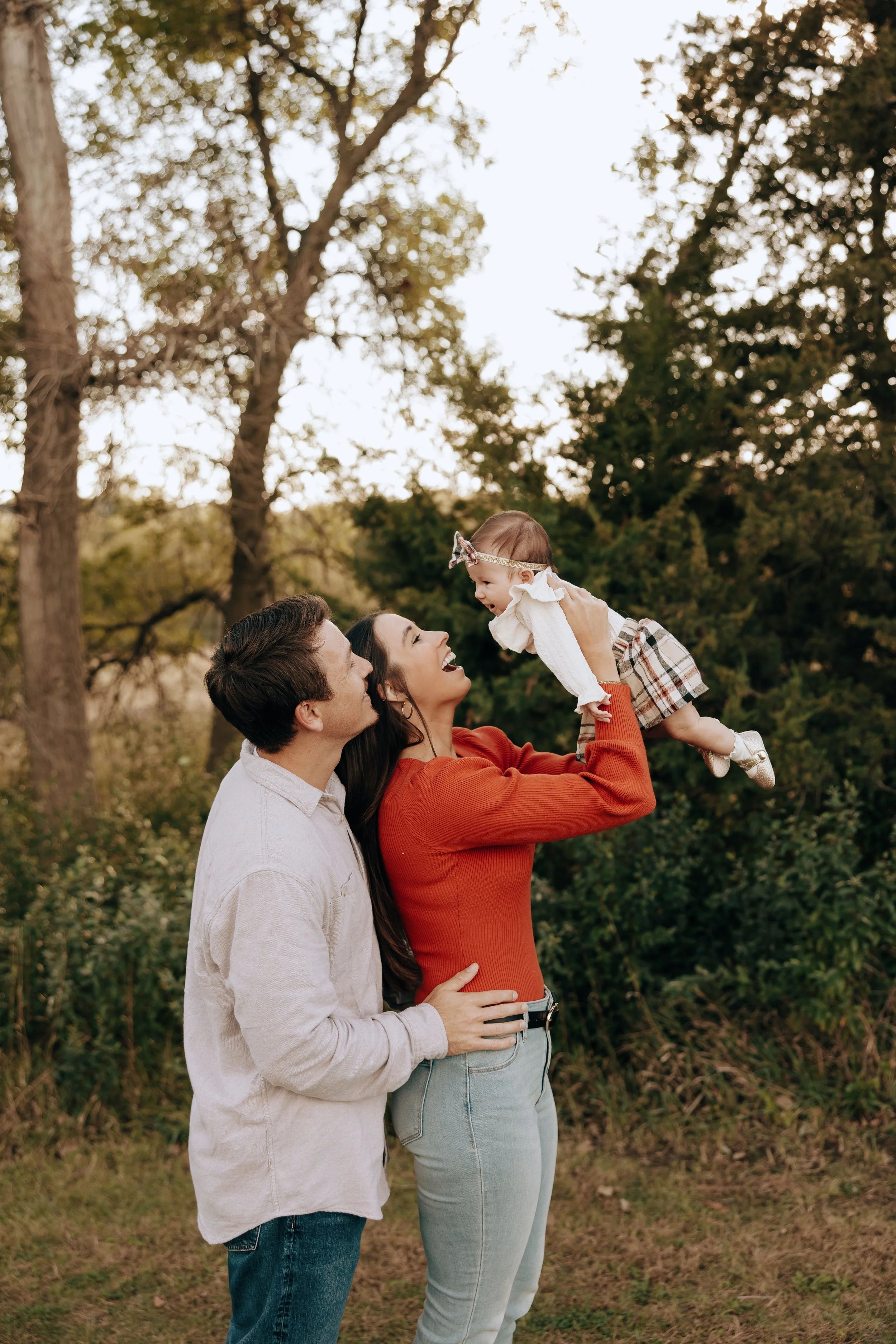 A happy family of three - a woman, a man, and a baby girl - outdoors in a wooded area, with the woman lifting the baby into the air as they all smile and enjoy the moment.