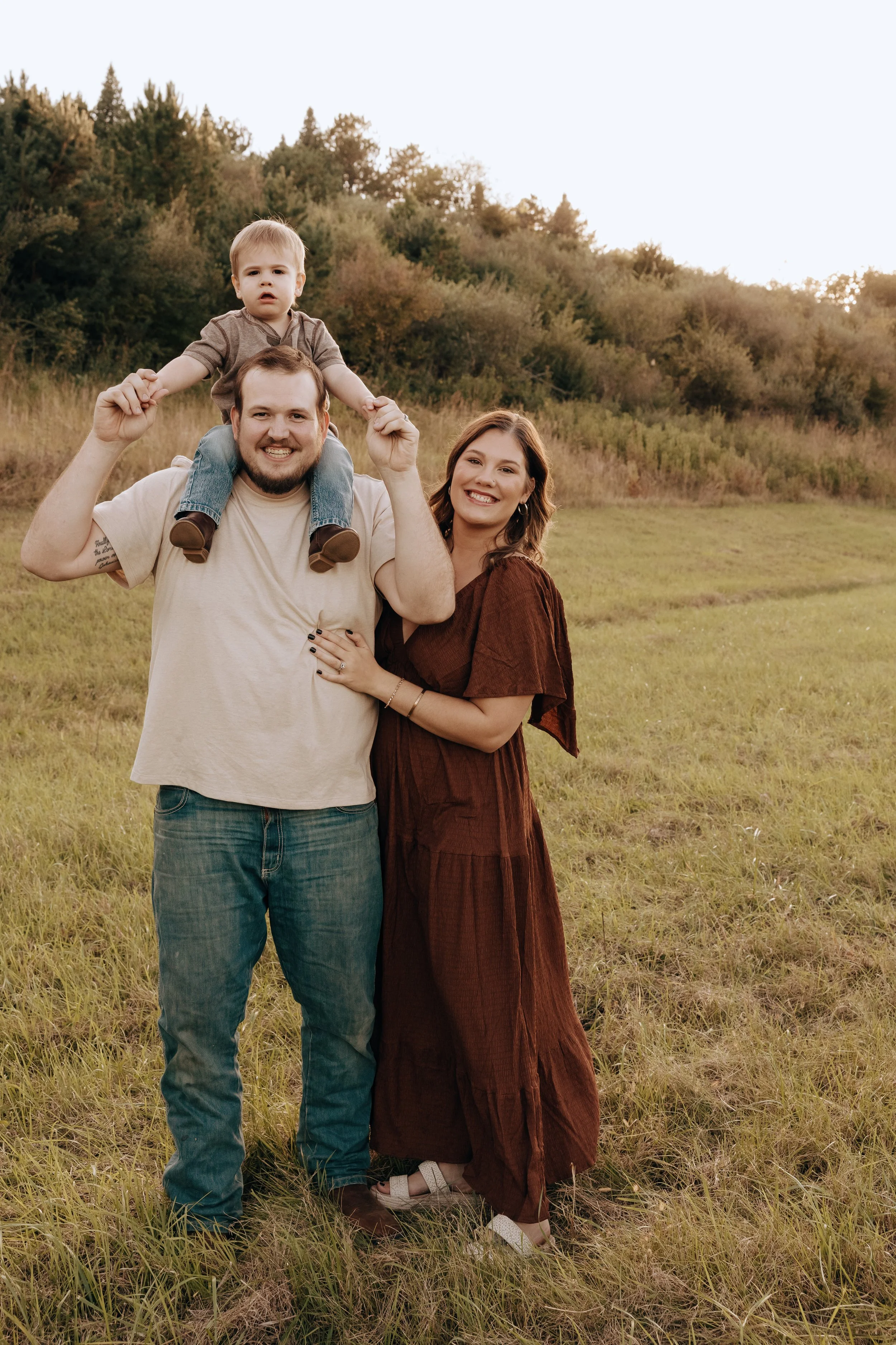 A happy family of three standing in a grassy field with trees in the background, the man carrying a young boy on his shoulders, both smiling and enjoying a day outdoors.