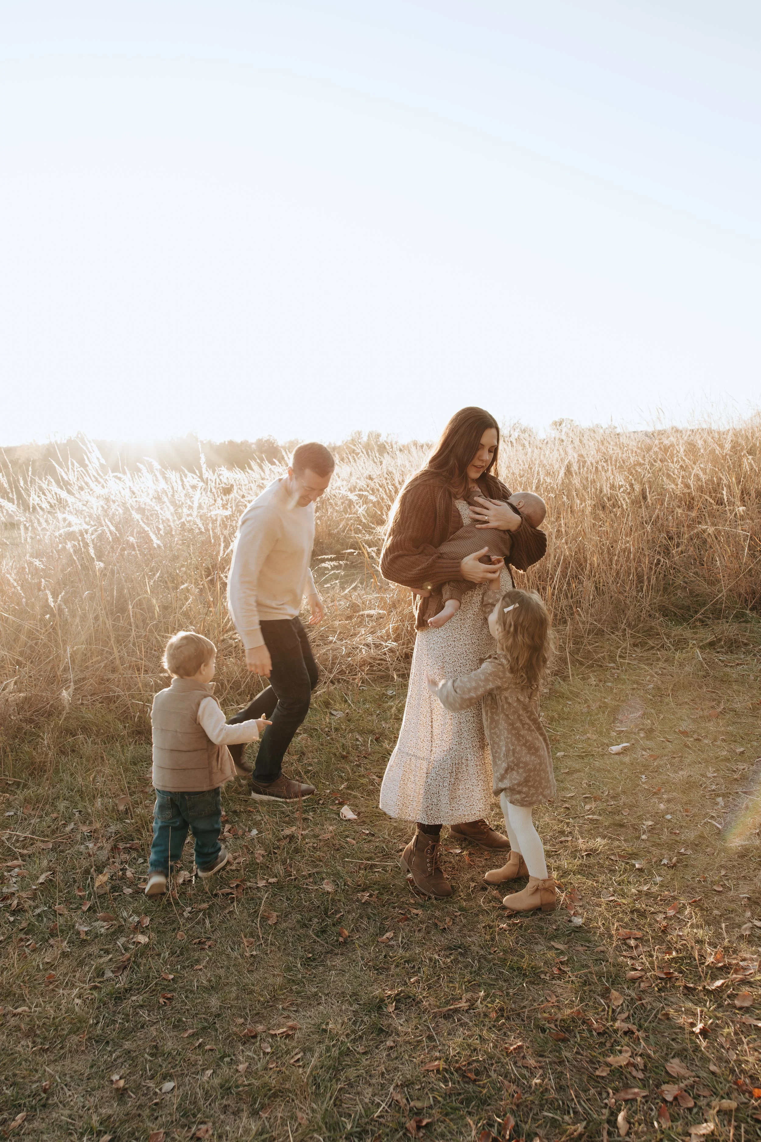 Omaha Nebraska Lifestyle Family Photographer. Katelyn Rowan Photography Omaha Nebraska. A woman breastfeeding a baby outside in a grassy field with sunset in the background, surrounded by three children.
