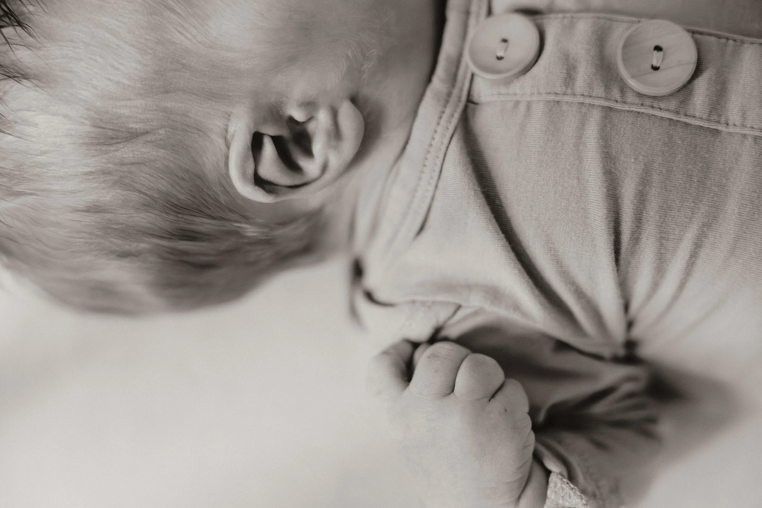 Newborn and Family Photography. Katelyn Rowan Photography. Omaha Nebraska newborn and family photographer. A baby with light hair is sleeping, wearing a shirt with buttons, and has their hand near their face.