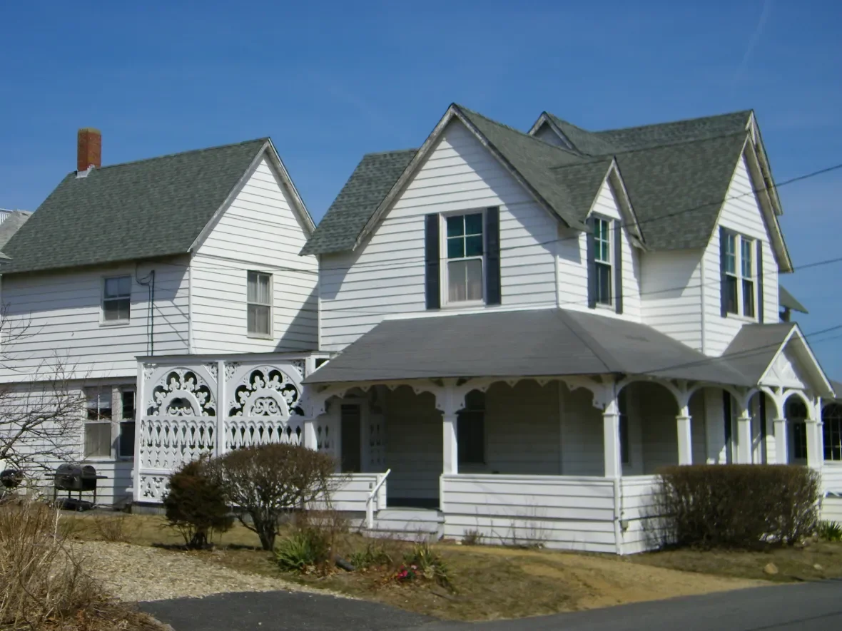 White two-story house with a porch and decorative trim, dark roofs, and a chimney under a blue sky.