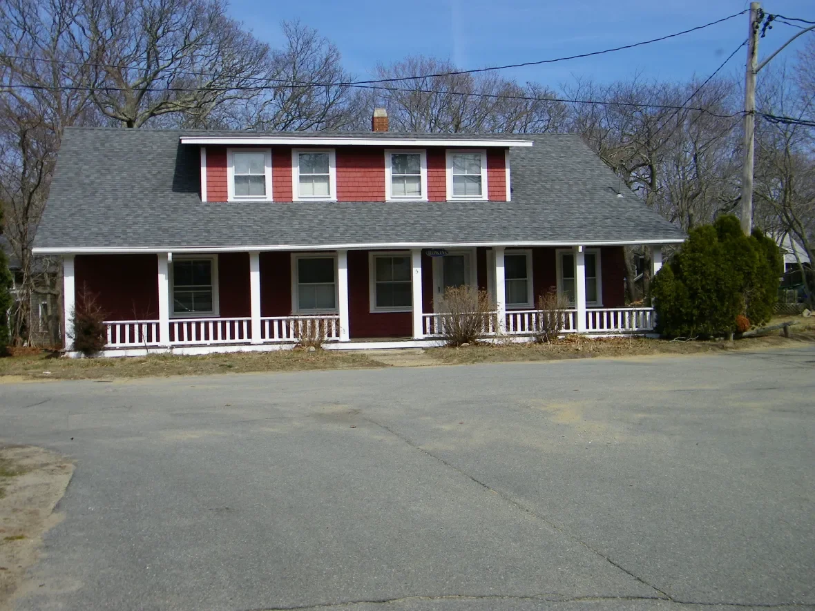 A two-story red house with a gray roof, white porch railing, and multiple windows, located in a residential area with bare trees and a paved street in the foreground.