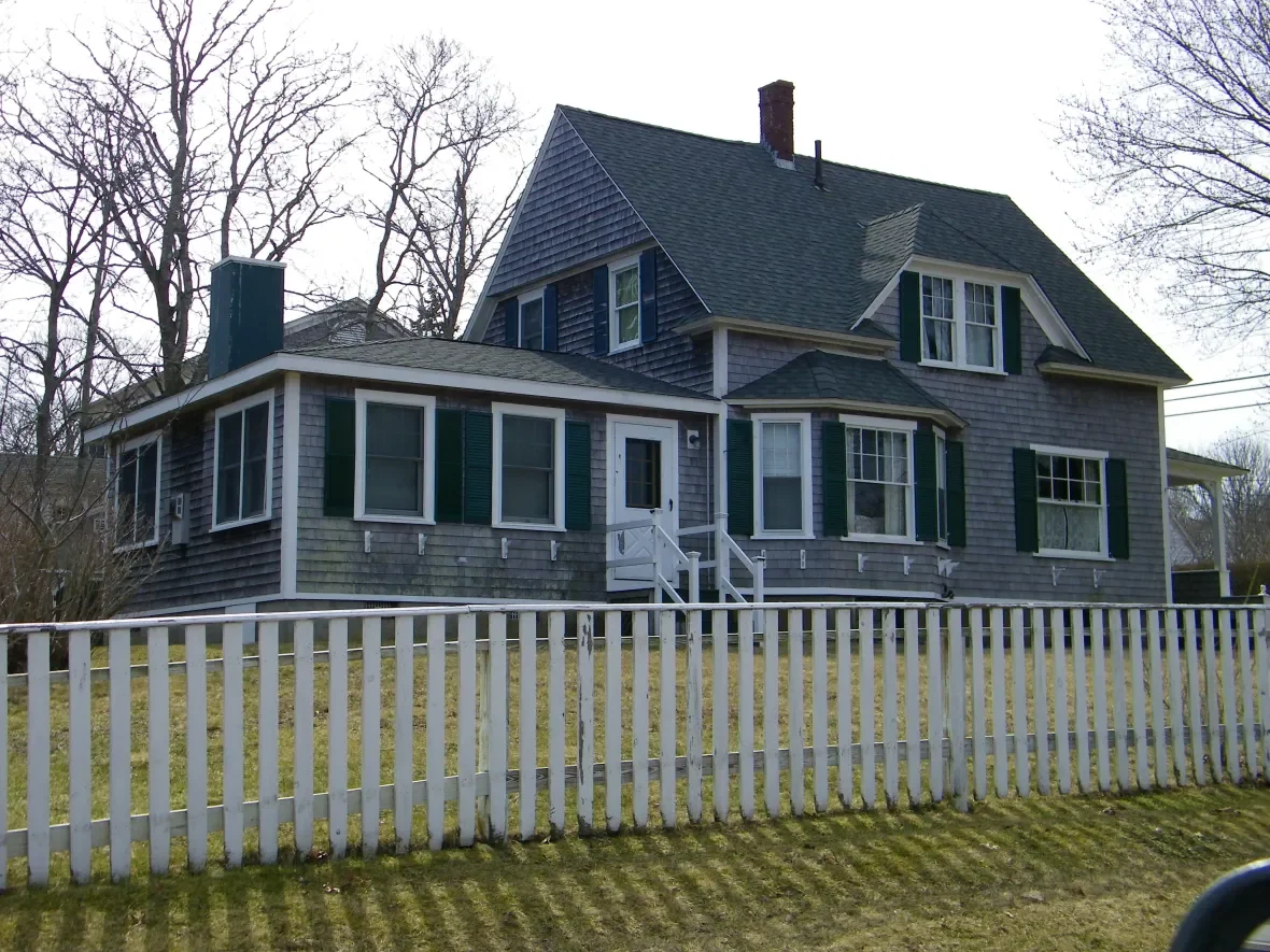 A large, gray, two-story house with green shutters, surrounded by a white picket fence, leafless trees in the background, and a cloudy sky.