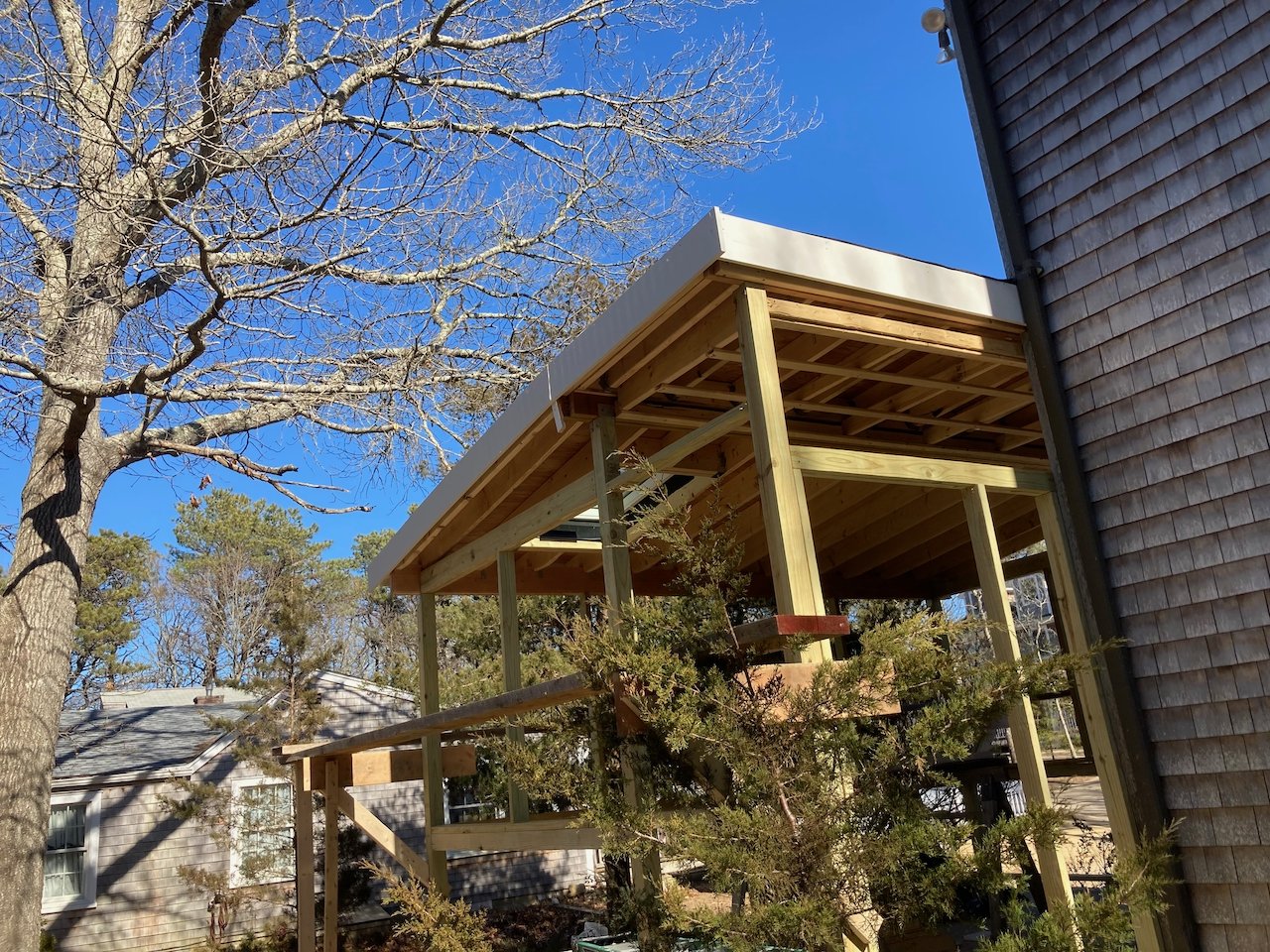 Construction of a wooden deck or balcony extension on the side of a house, with the framework in place and no railing yet, surrounded by trees with bare branches.
