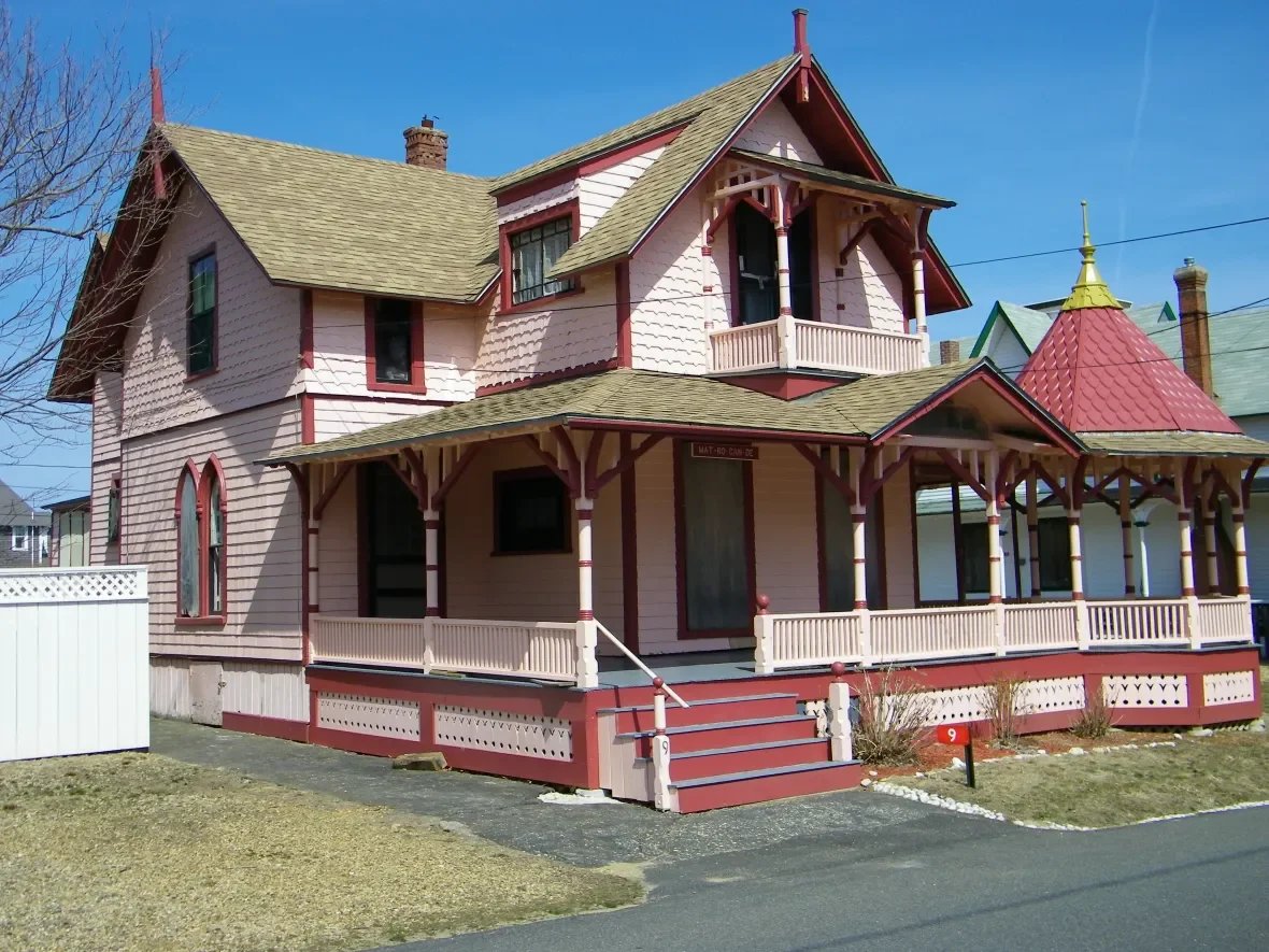 Pink Victorian house with red trim and a wraparound porch, featuring a turret with a red conical roof, under a clear blue sky.