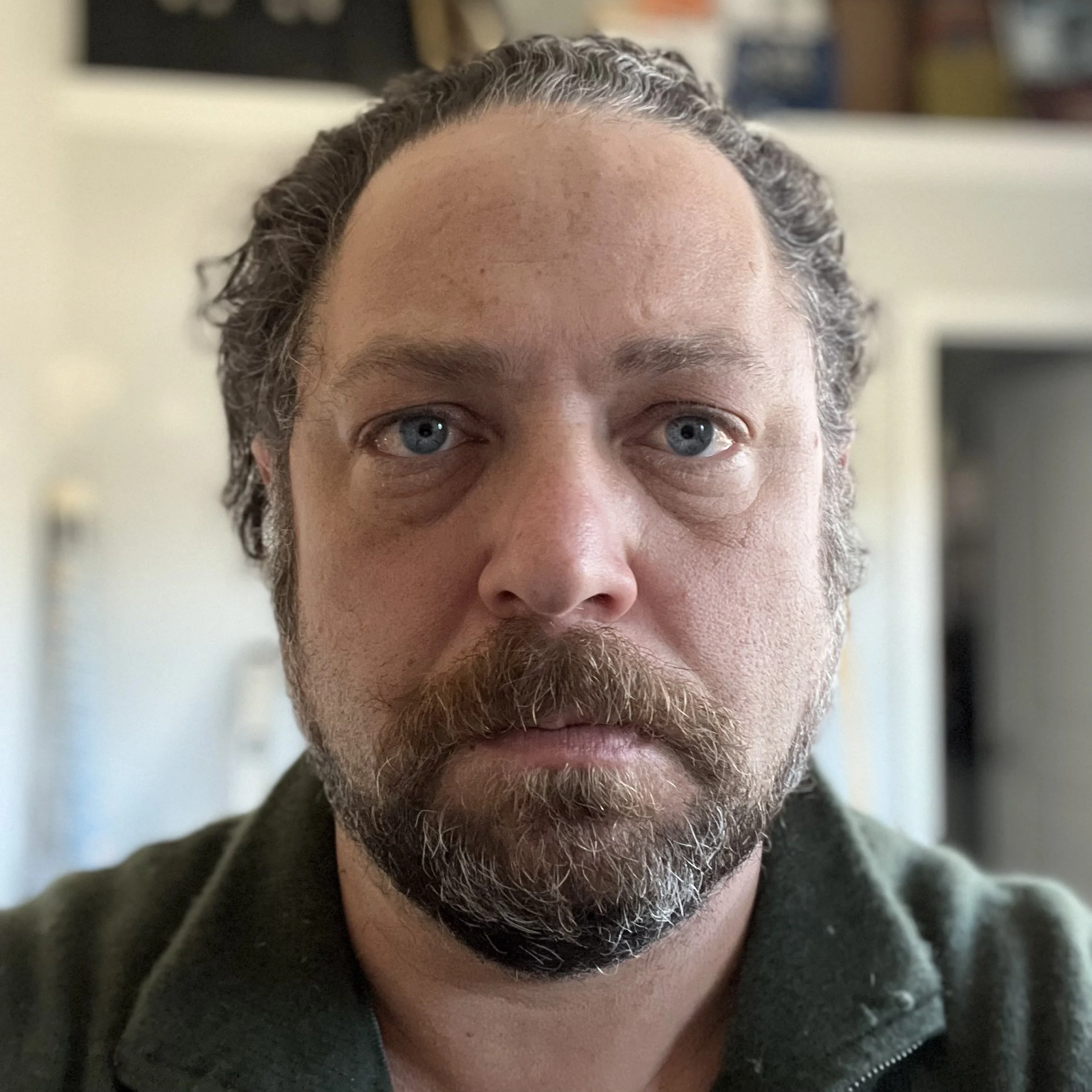 Close-up of a man with blue eyes, dark curly hair, and a beard, looking directly at the camera with a neutral expression, indoors with a blurred background.