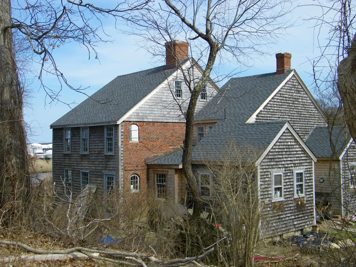 A large old house with grey shingles, brick chimney, and multiple windows, surrounded by leafless trees and overgrown bushes.