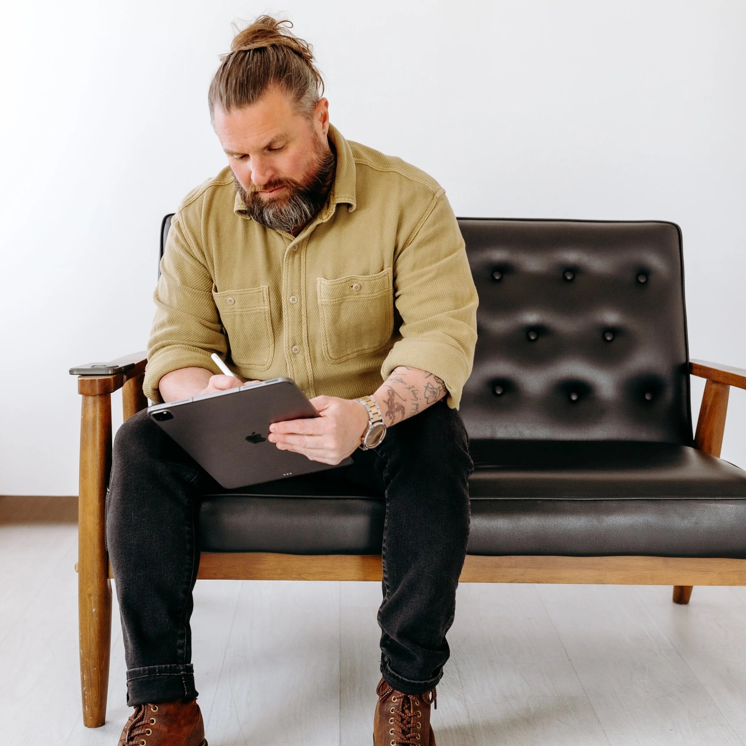 Man with a beard and tattoo on his arm sitting on a wooden and leather black bench, writing on a digital tablet with a stylus.