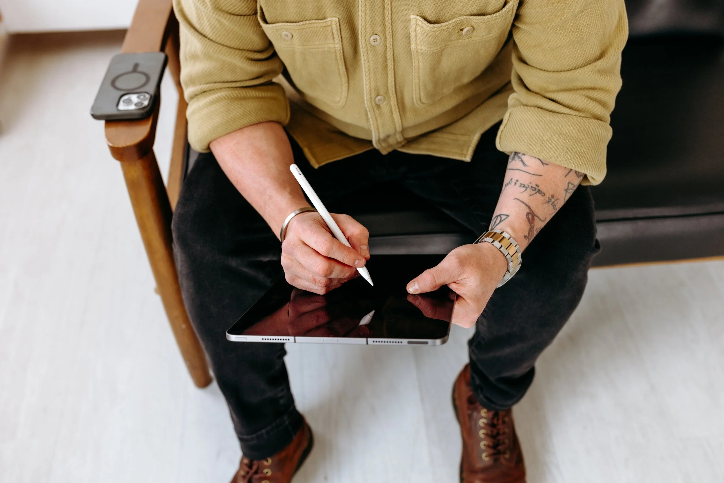 A person sitting on a wooden chair using a stylus on a tablet, with a smartphone placed on the armrest of the chair.