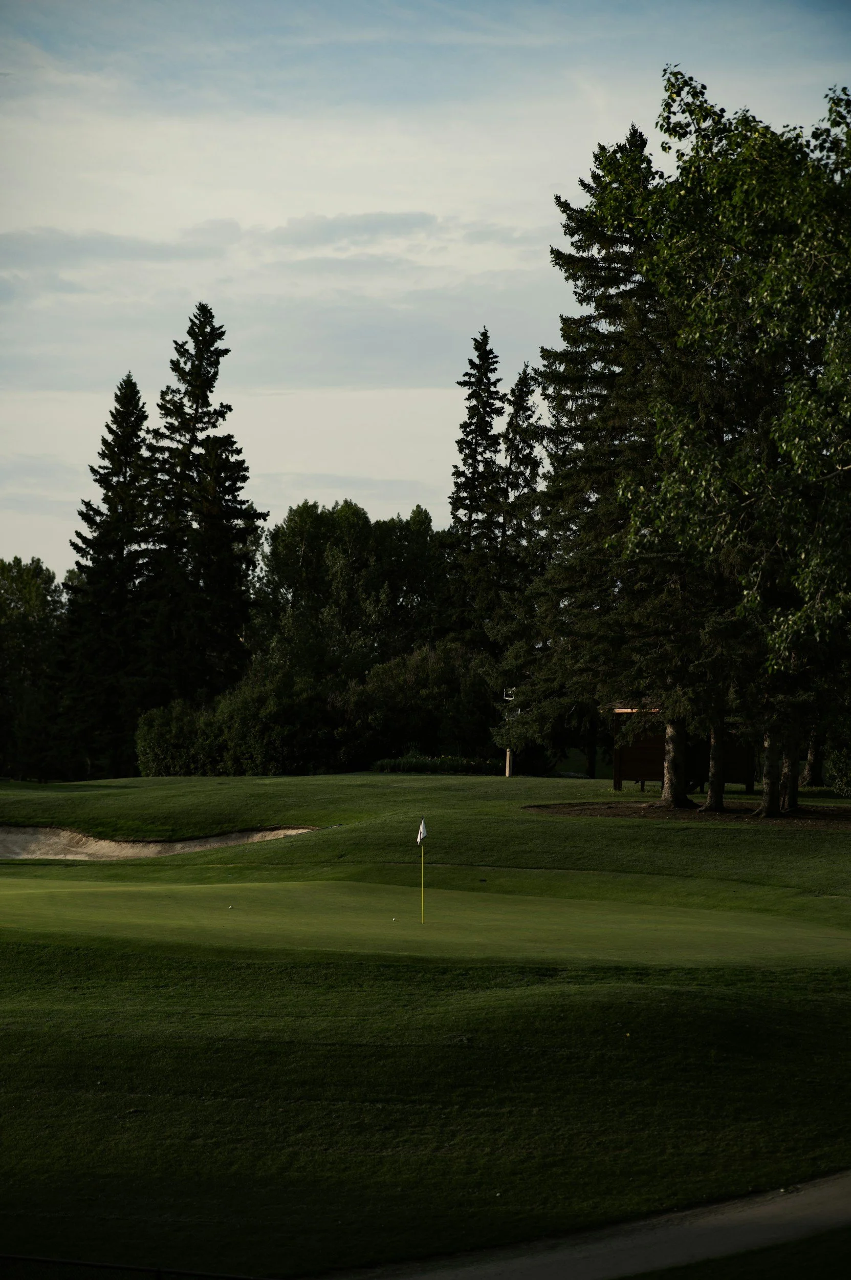 A golf course green with a flag, surrounded by trees and sand bunkers, under a cloudy sky.