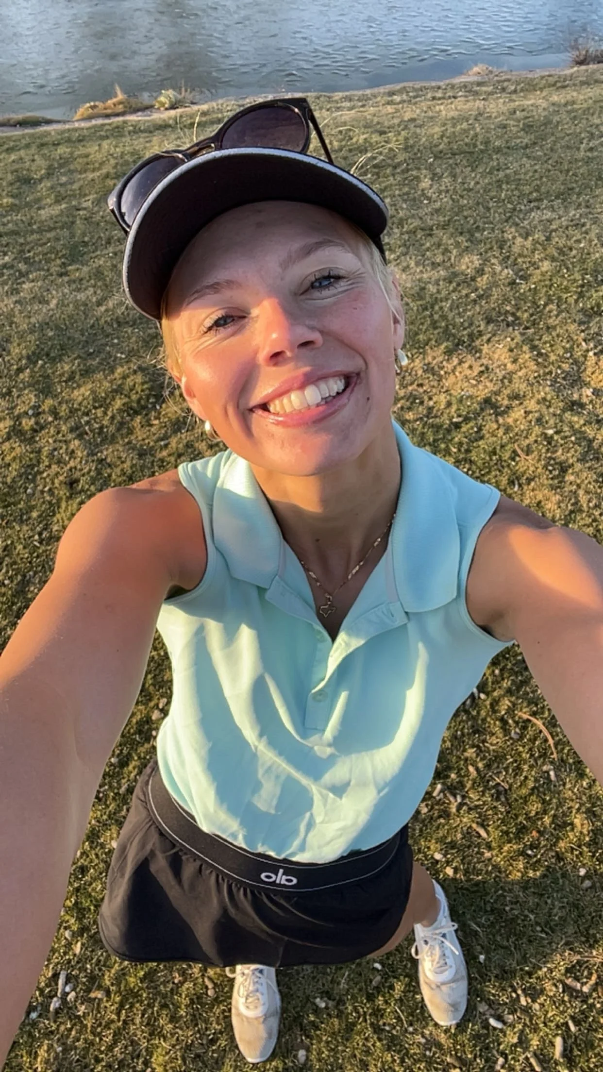 A smiling woman takes a selfie outdoors near a body of water during sunset, wearing a light green sleeveless polo shirt, black athletic skirt with a brand logo, white sneakers, a baseball cap with sunglasses on top, and a necklace. Brianna Spencer.
