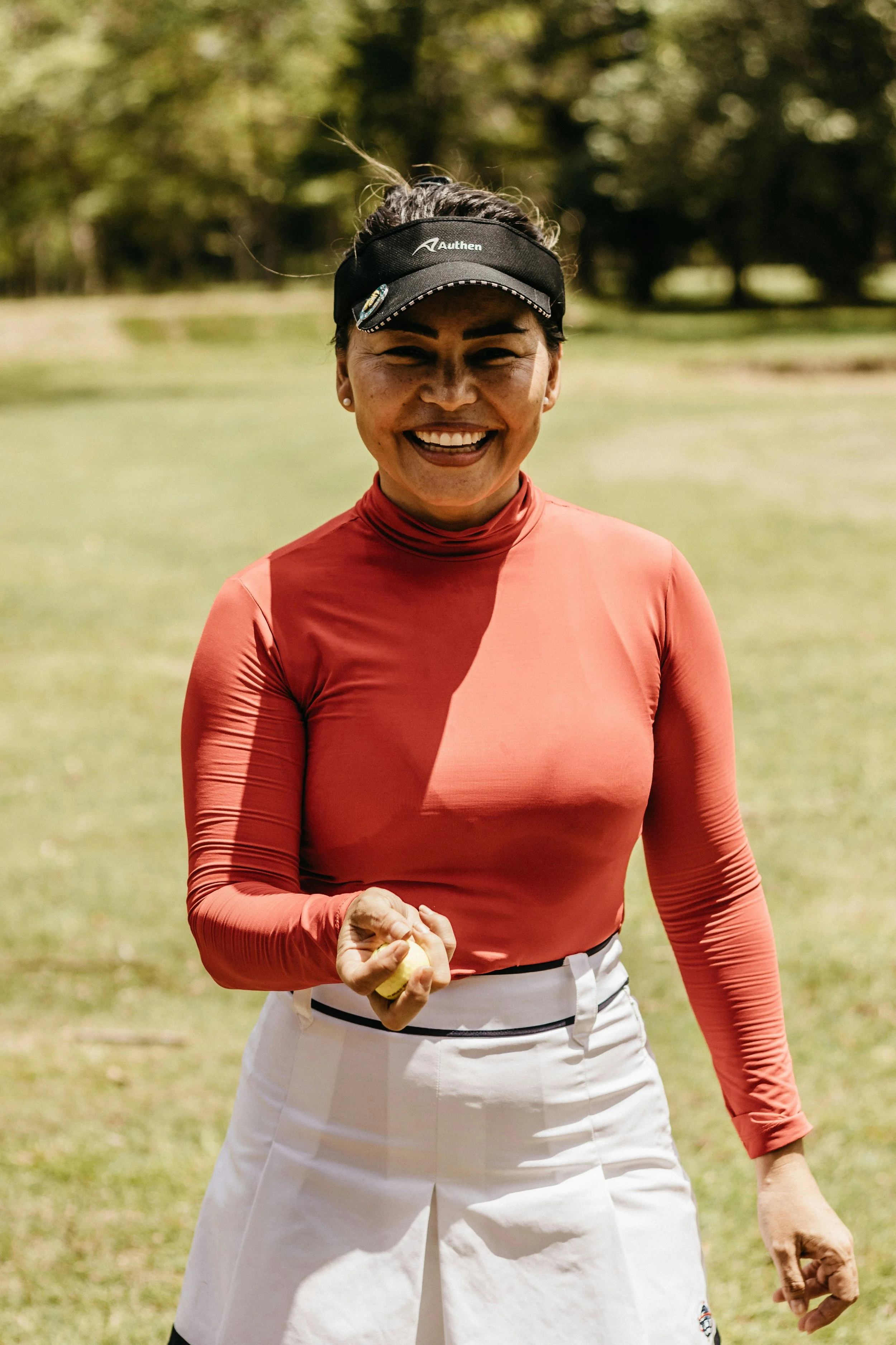 A woman smiling outdoors, holding a golf ball in her right hand, wearing a black cap, red long-sleeve shirt, and white skirt, with green trees and grass in the background.
