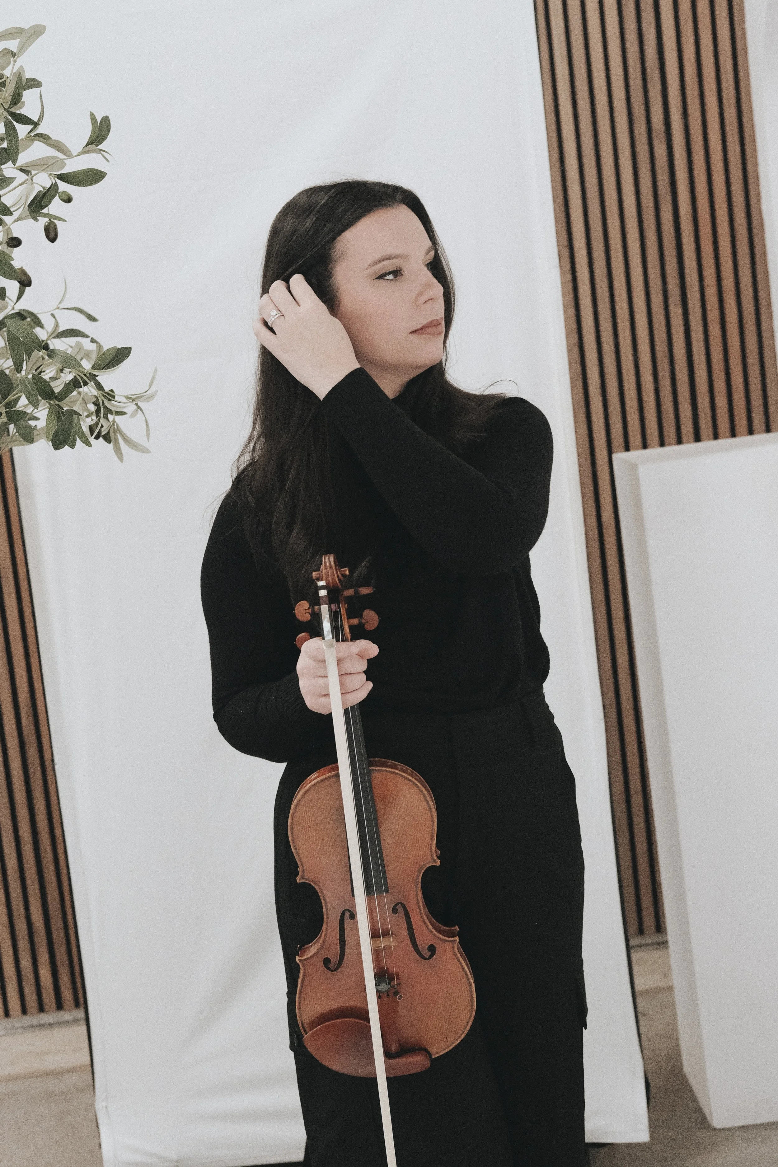 A woman with dark hair wearing a black long sleeve top, holding a violin and adjusting her hair, standing in a minimalist room with wooden accents and a plant.