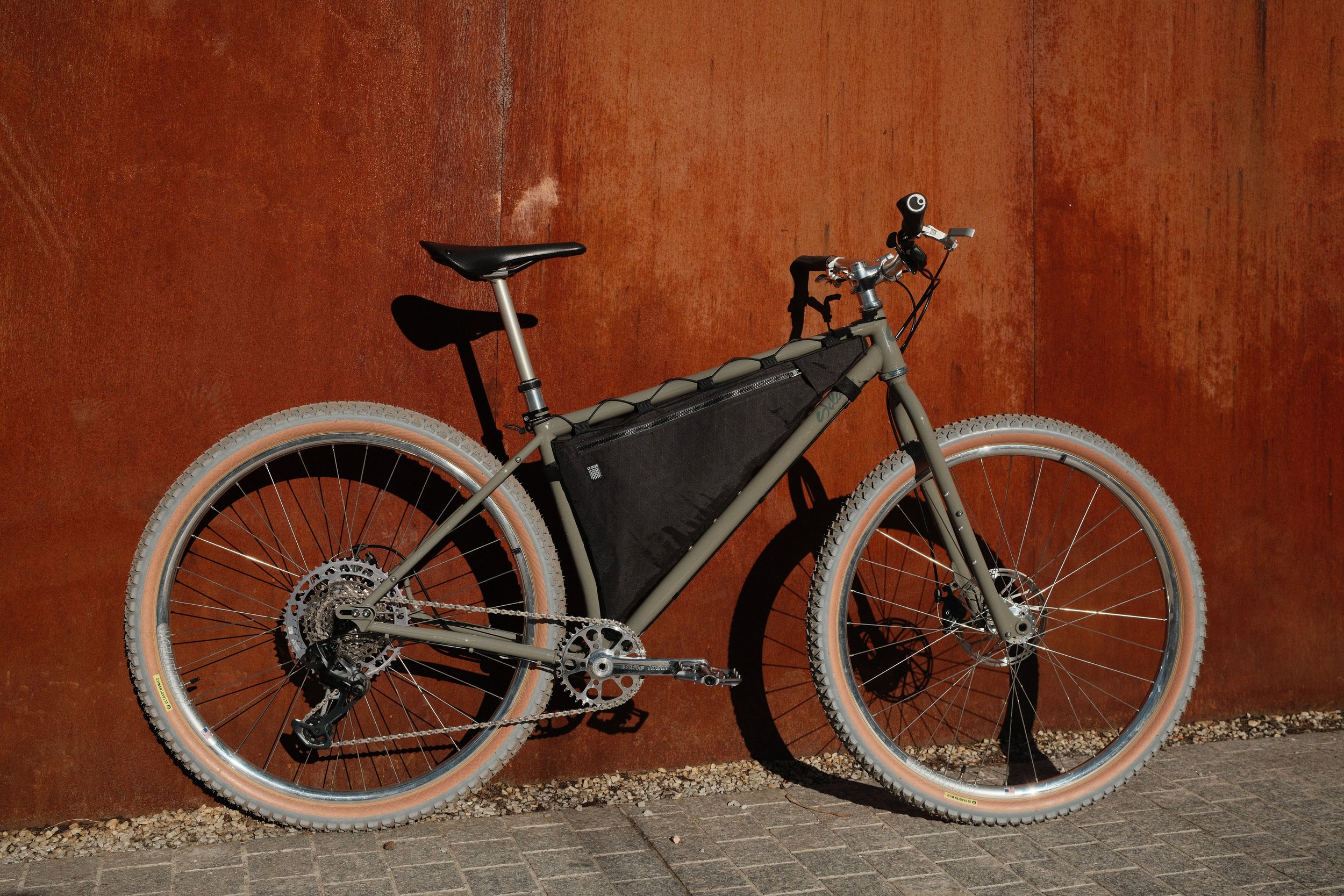 Gray mountain bike with fat tires, black saddle, and a large black battery pack mounted on the frame, leaning against a rusty red wall.