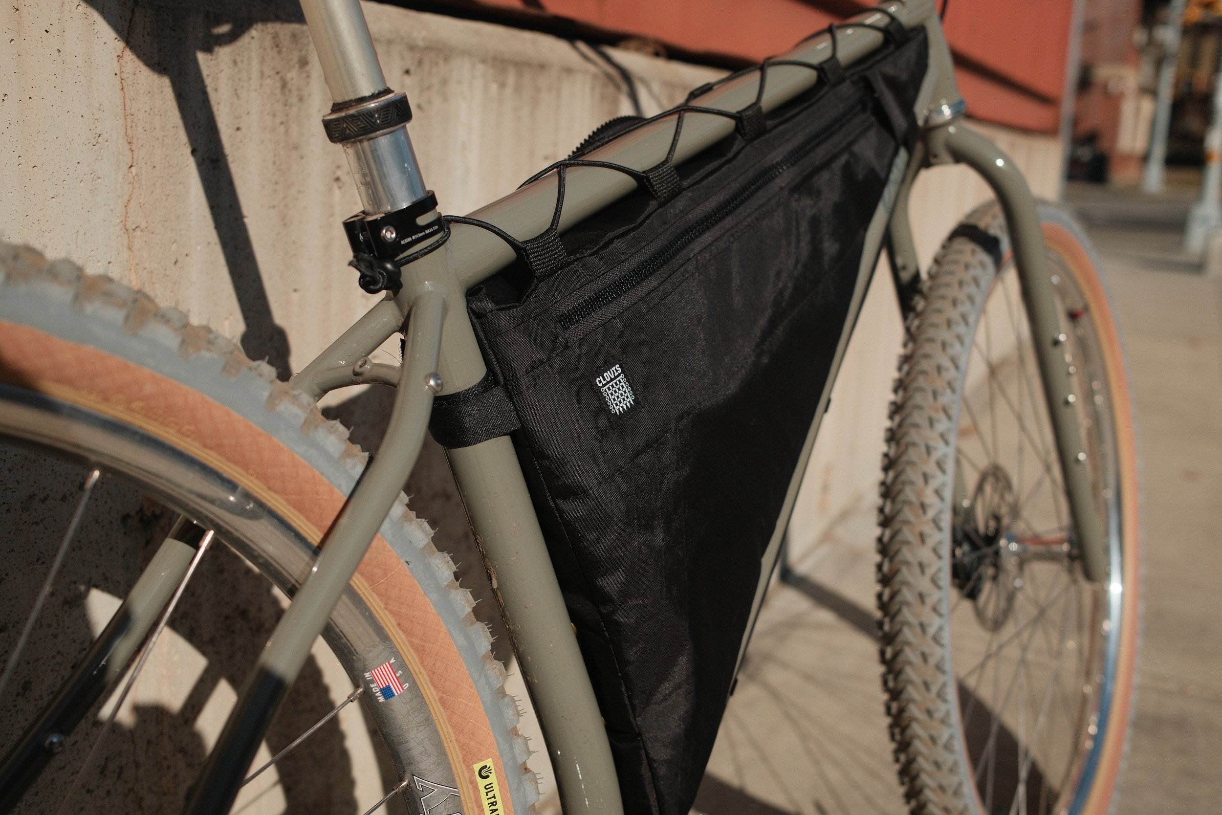 Close-up of a beige mountain bike with a black frame bag attached to the top tube, leaning against a concrete wall on a sunny day.
