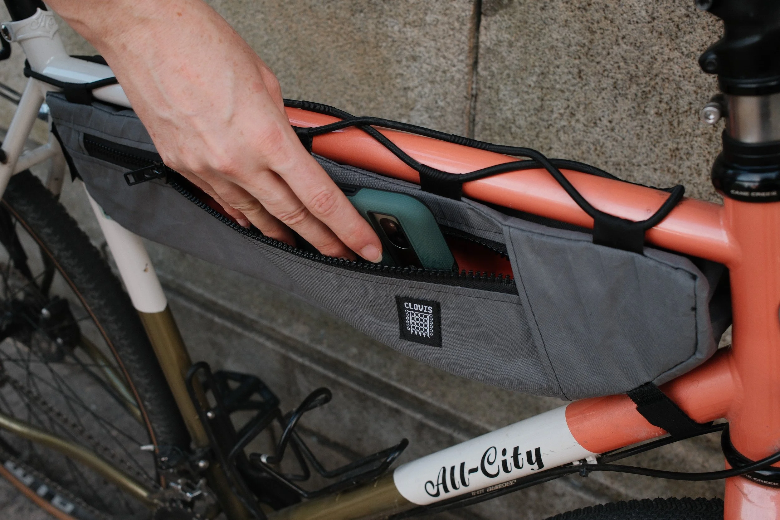 A person places a smartphone into a gray bicycle top tube bag attached to a bike with a pink frame that has the words "All-City" on it. The bag is secured with black straps and has a zippered pocket.
