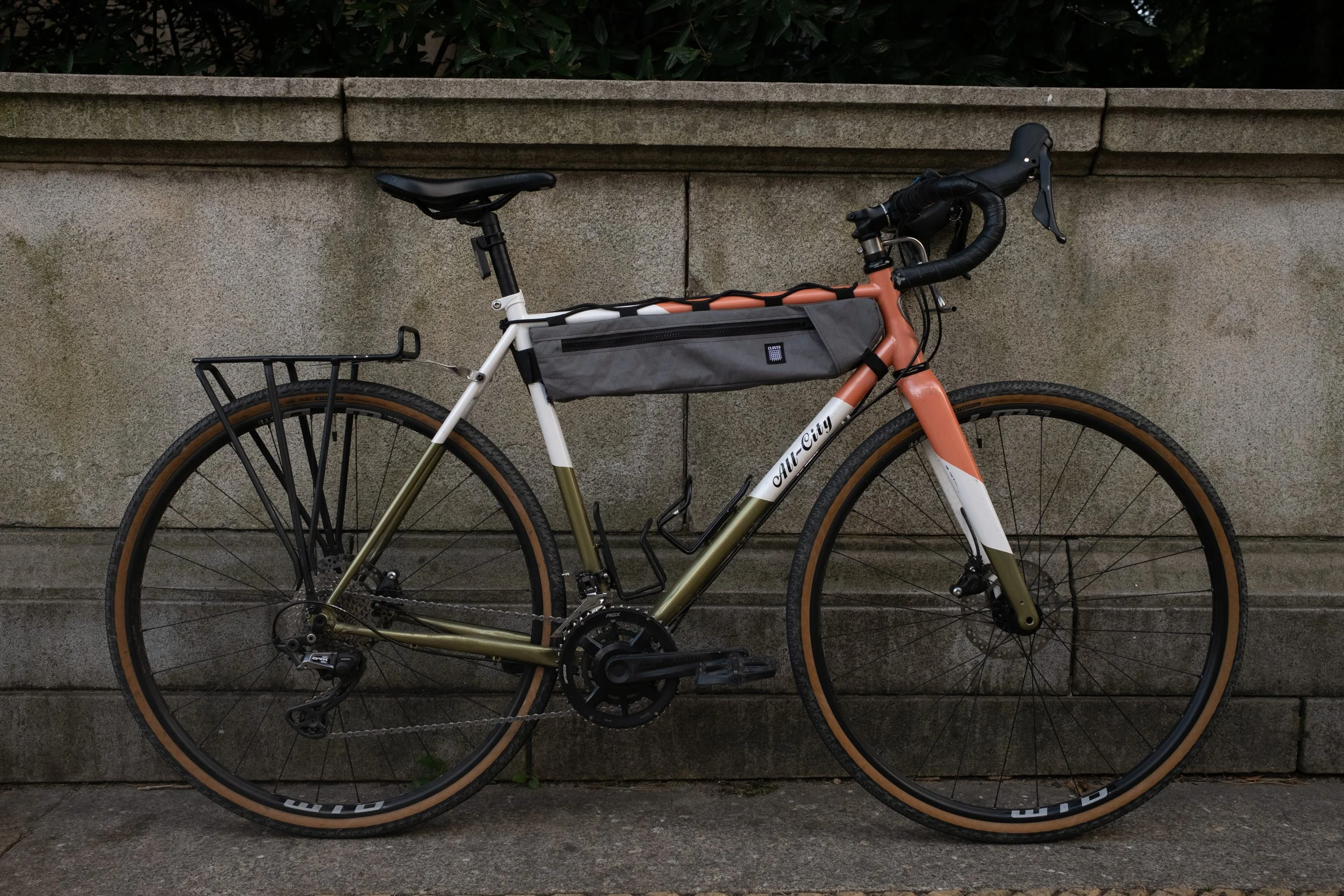 A modern road bicycle with a frame in shades of pink, white, and olive green, featuring a gray storage bag mounted on top tube, black handlebars with handlebar tape, and large tires with tan sidewalls, leaning against a concrete wall outdoors.