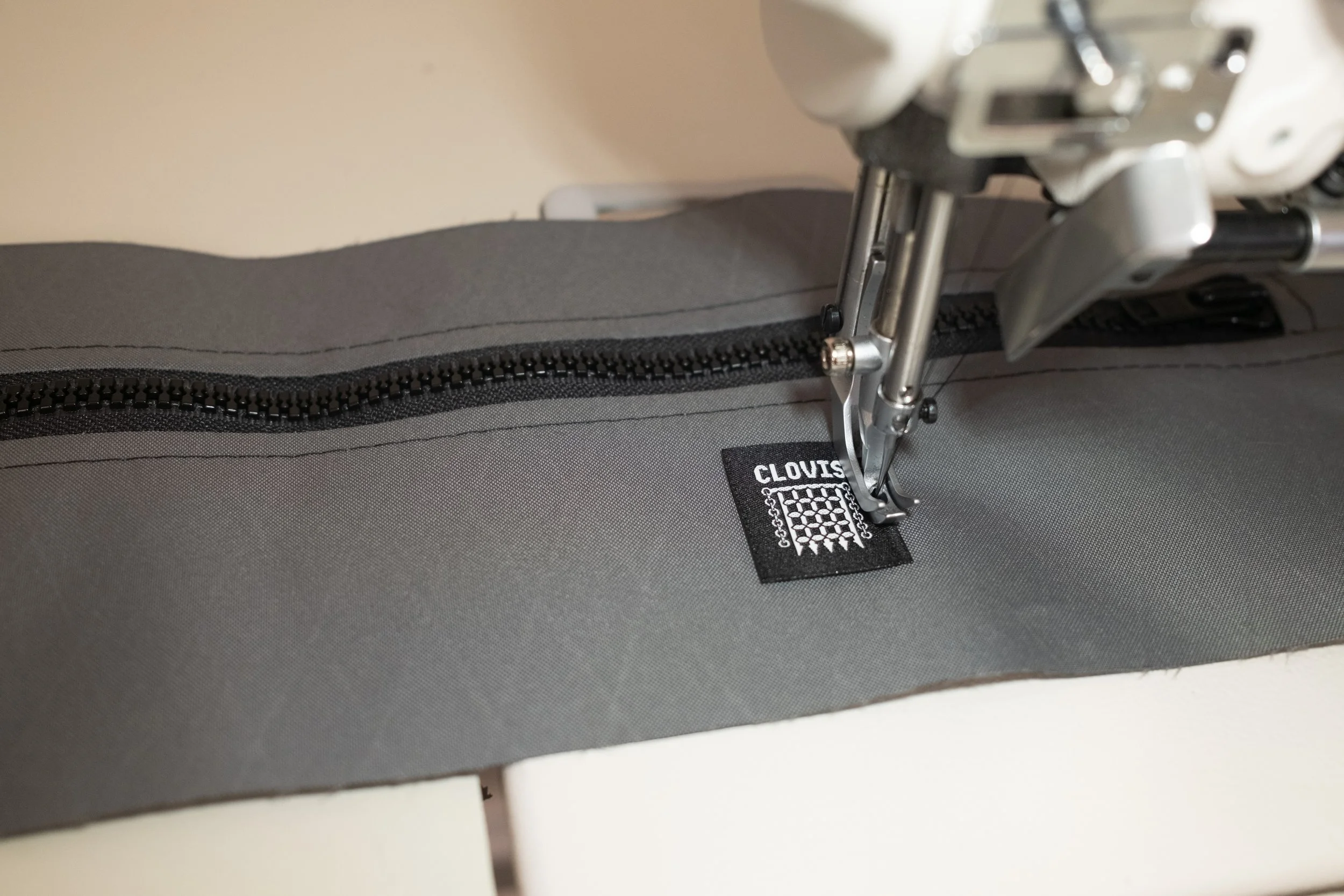 Close-up of a sewing machine stitching a black label with white text 'CLOVIS' onto gray fabric with a zipper.