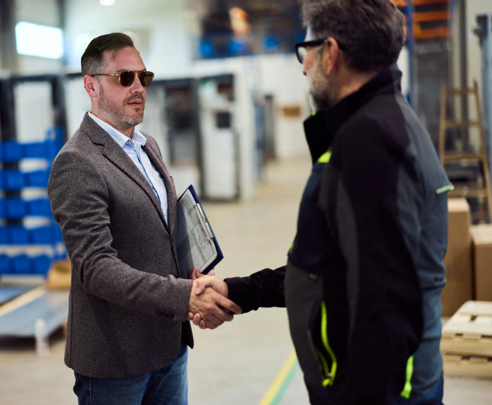 Two men shaking hands in a warehouse, one holding a clipboard and wearing sunglasses, the other dressed in black with yellow accents.