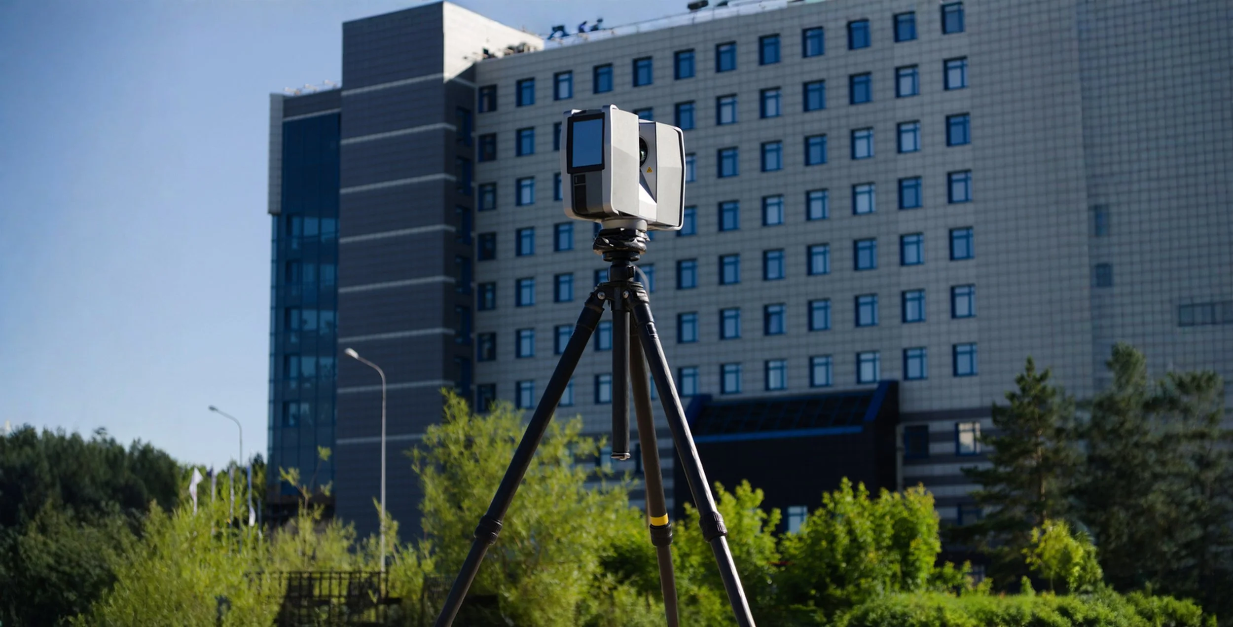 A 3D scanner mounted on a tripod outdoors in front of a modern multi-story building with many windows, surrounded by green trees and bushes.