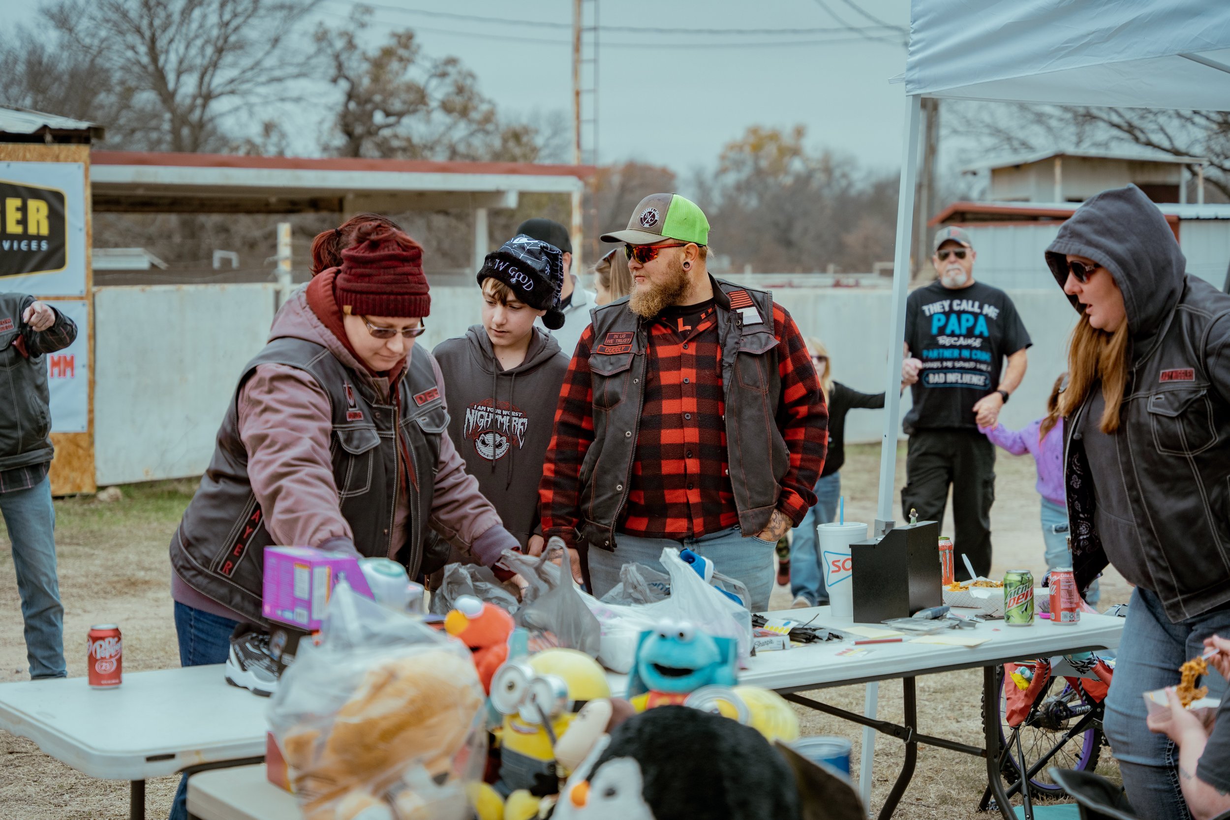 Group of people at an outdoor event, gathered around a table with toys, snacks, and soda cans.