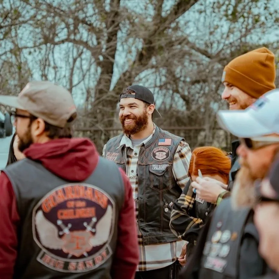 Group of people gathered outdoors, some wearing leather vests with patches, possibly motorcycle club members, amidst autumn trees.