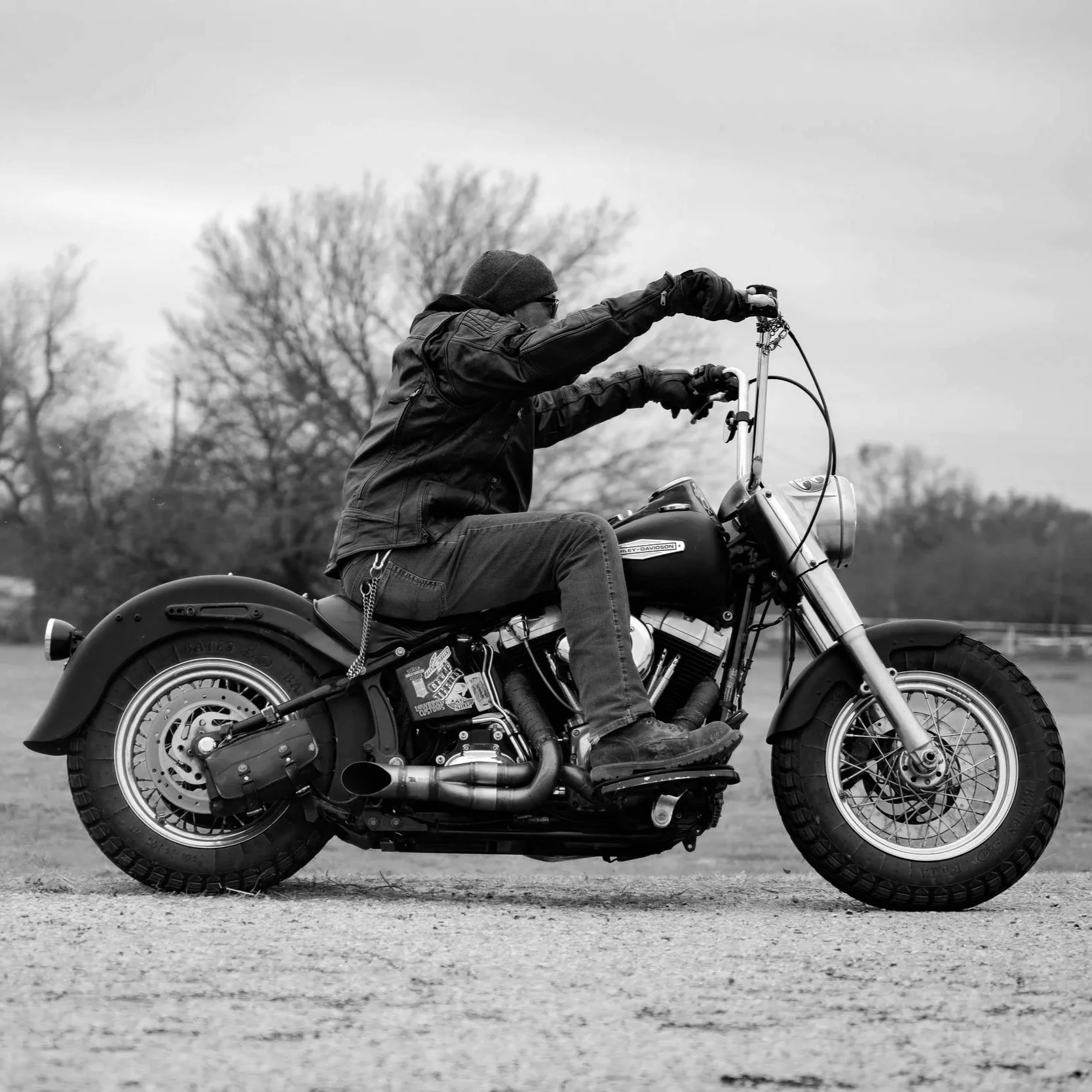 A man riding a motorcycle outdoors on a cloudy day, dressed in a leather jacket, jeans, gloves, a beanie, and sunglasses, with trees in the background.