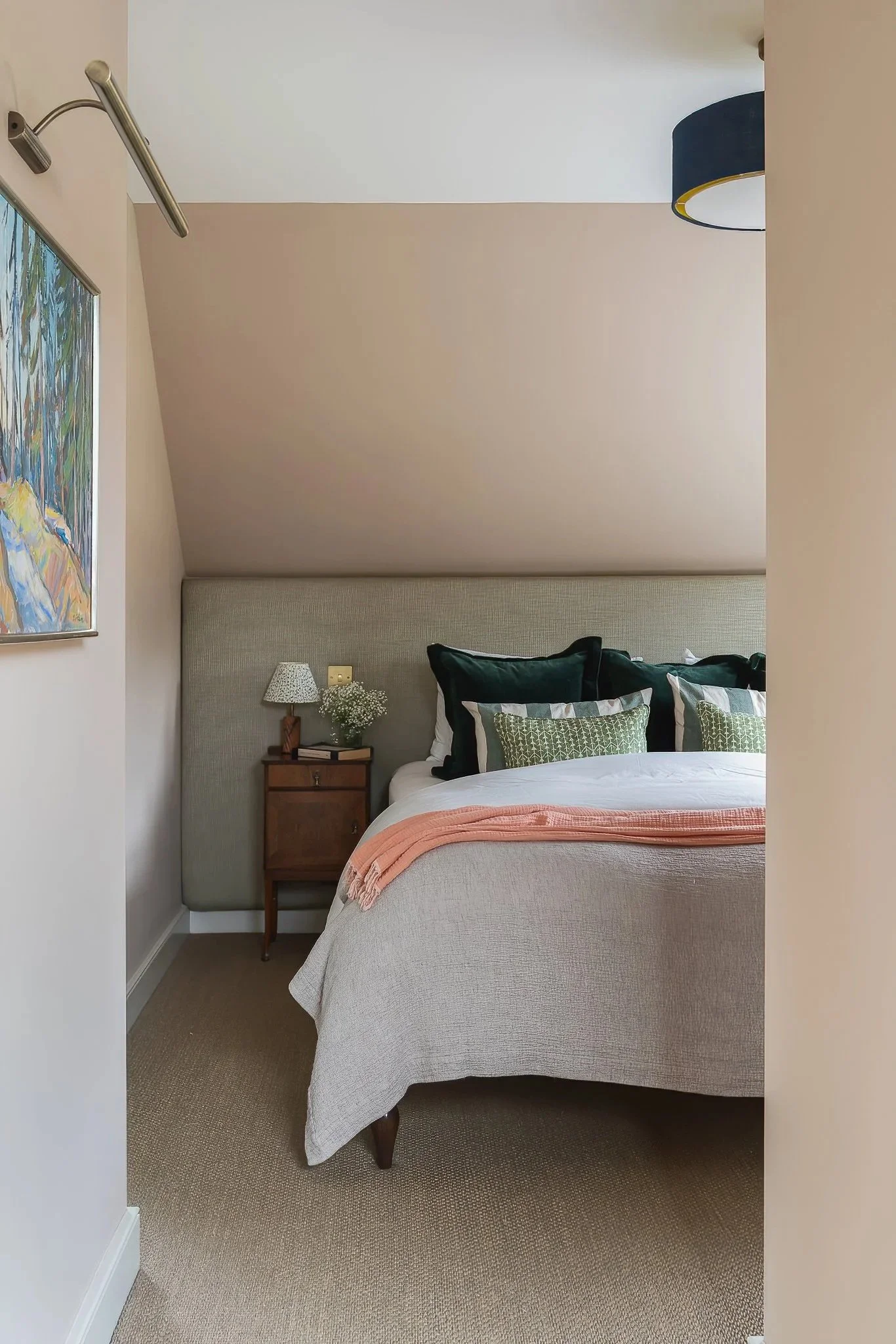 A cozy bedroom with a bed that has a light beige upholstered headboard, green and white pillows, a peach throw blanket, and a beige bedspread. A small wooden nightstand with a lamp, a book, and a small vase with flowers is beside the bed. The room ha