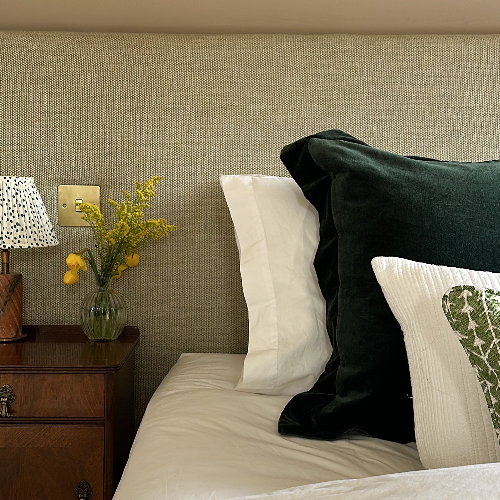 A bedroom scene with a bedsheet, pillows, a nightstand with a vase of yellow flowers, and a table lamp, against a textured beige wall.