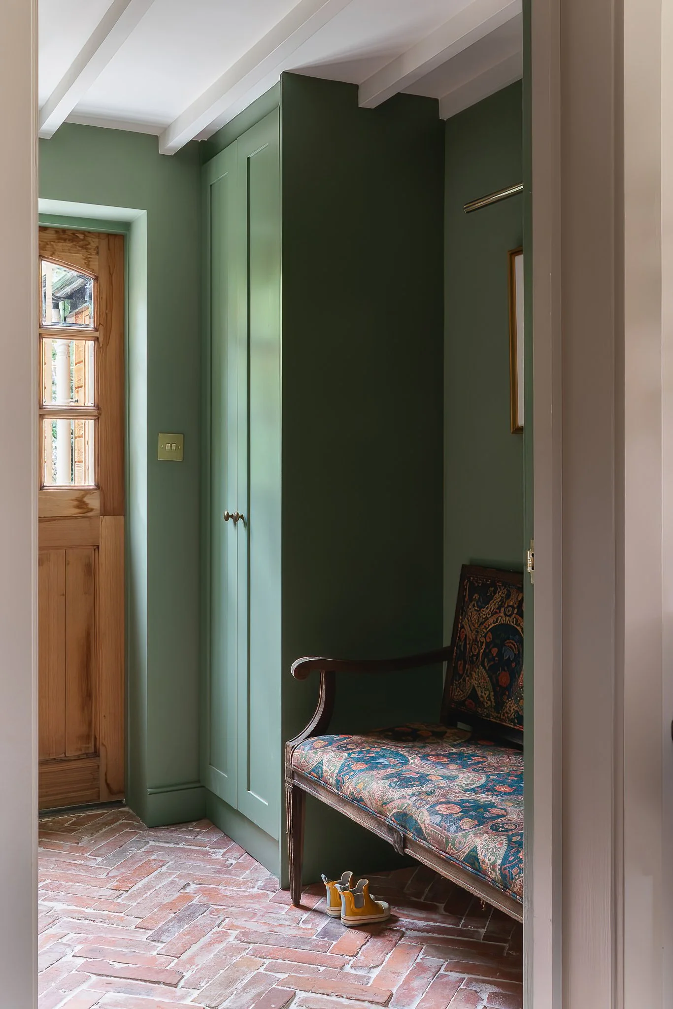 Interior view of an entryway with green walls, a wooden front door, a built-in green closet, a vintage upholstered bench with dark wood frame, and a pair of yellow and white shoes on the brick floor.