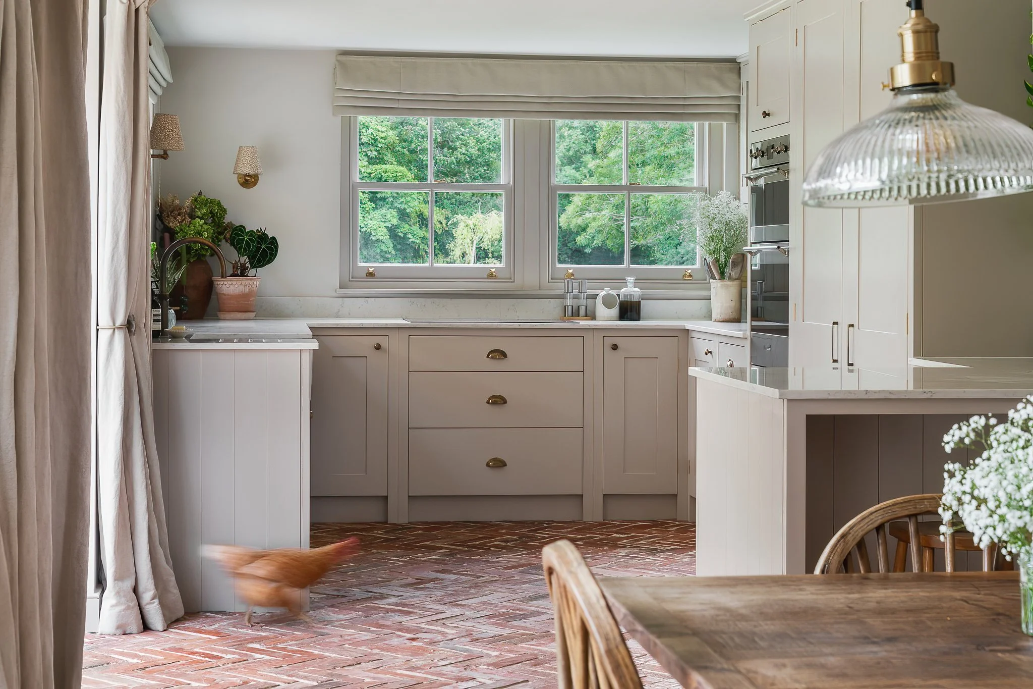 A bright kitchen with white cabinetry and a large window overlooking greenery. There are plants on the counter, a wooden dining table with a vase of white flowers, and a chicken walking on a brick floor.