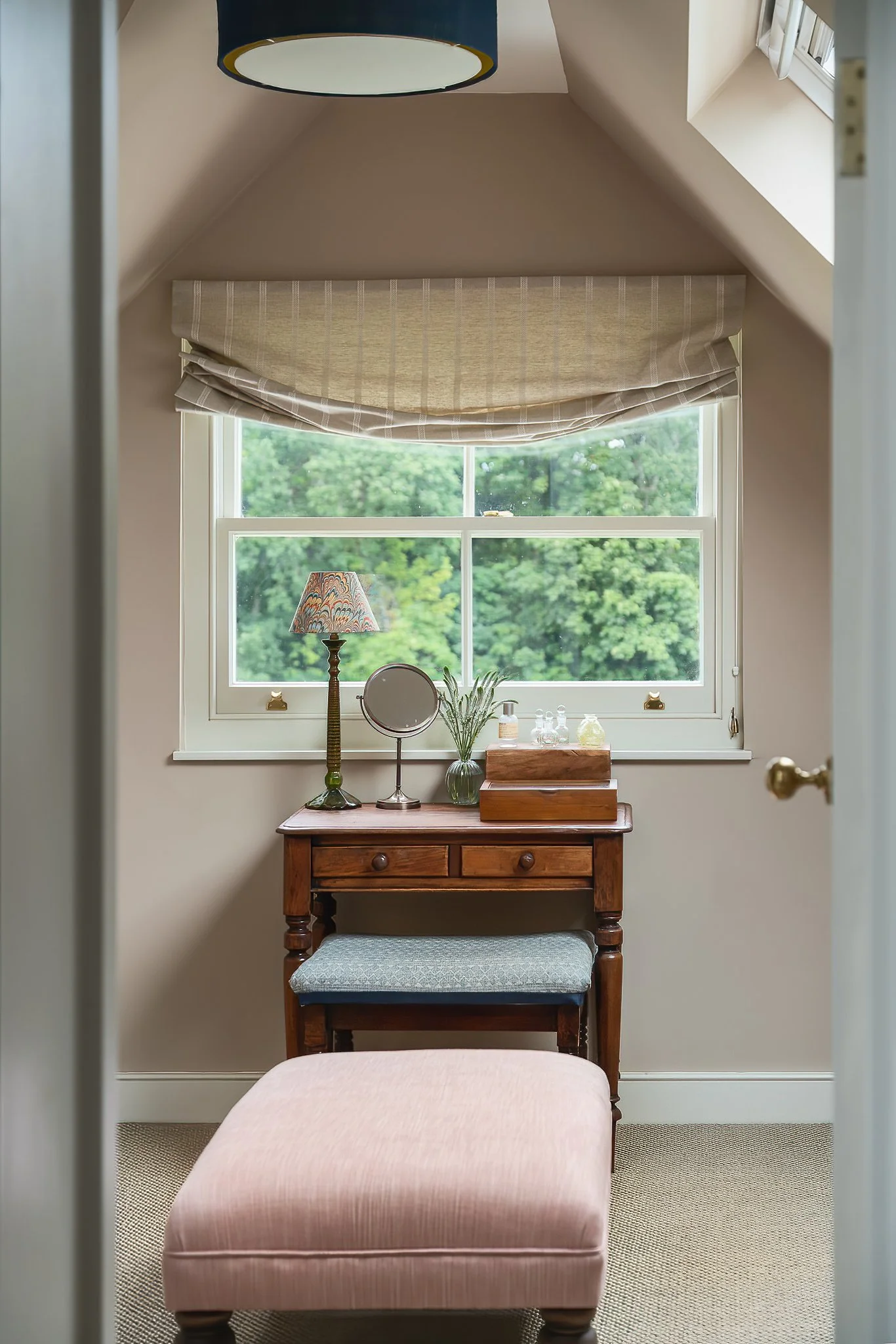 A small wooden vanity table with a cushioned stool in front, placed beneath a window with a beige Roman shade. The table holds a lamp, a mirror, a vase with lavender, and some glass bottles, with a view of green trees outside.