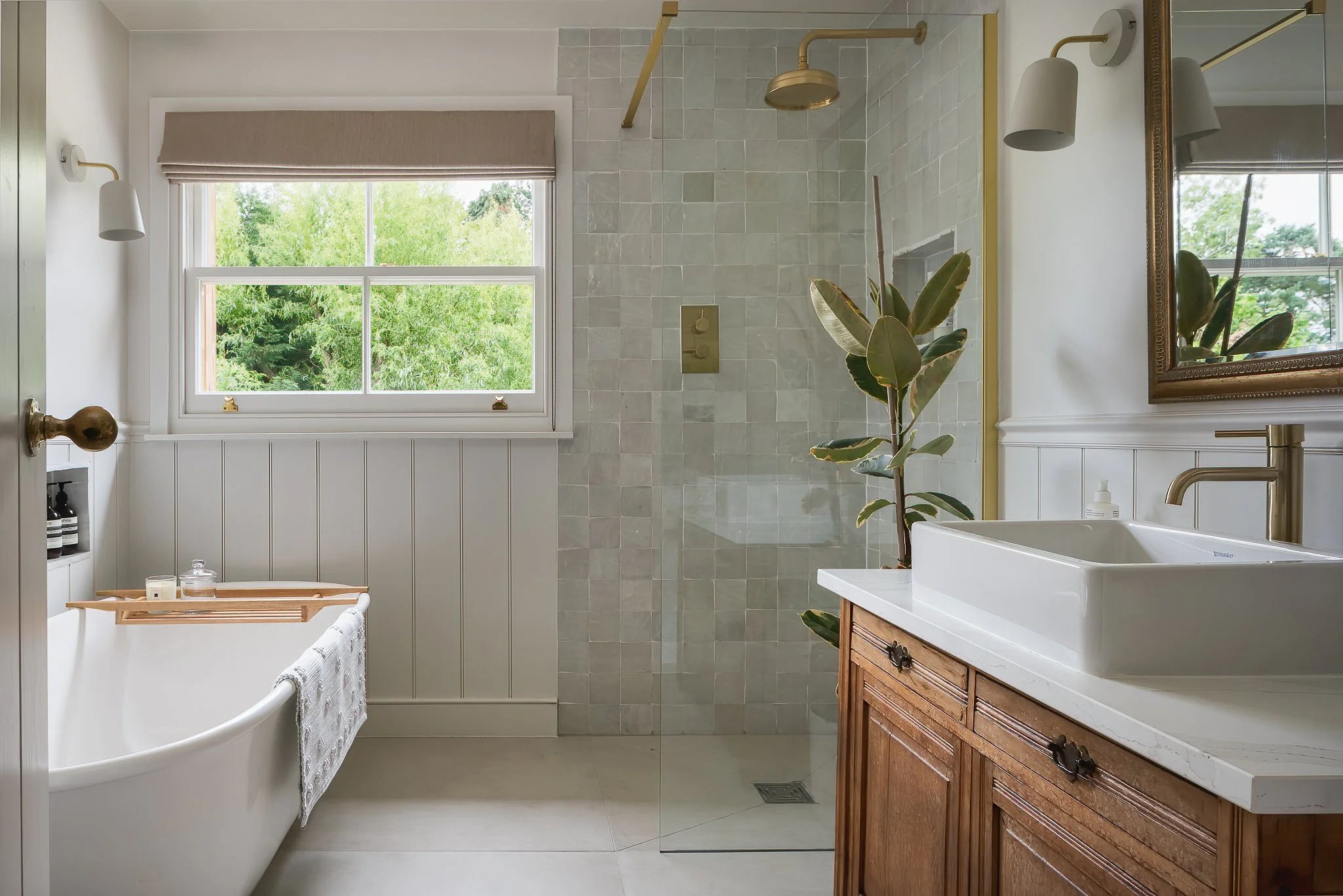 A bright bathroom featuring a large window with a view of greenery, a freestanding bathtub with a wooden tray and candles, a walk-in shower with beige tiles and a gold showerhead, a wooden vanity with a white vessel sink, a mirror with a gold frame, a potted plant, and neutral decor.
