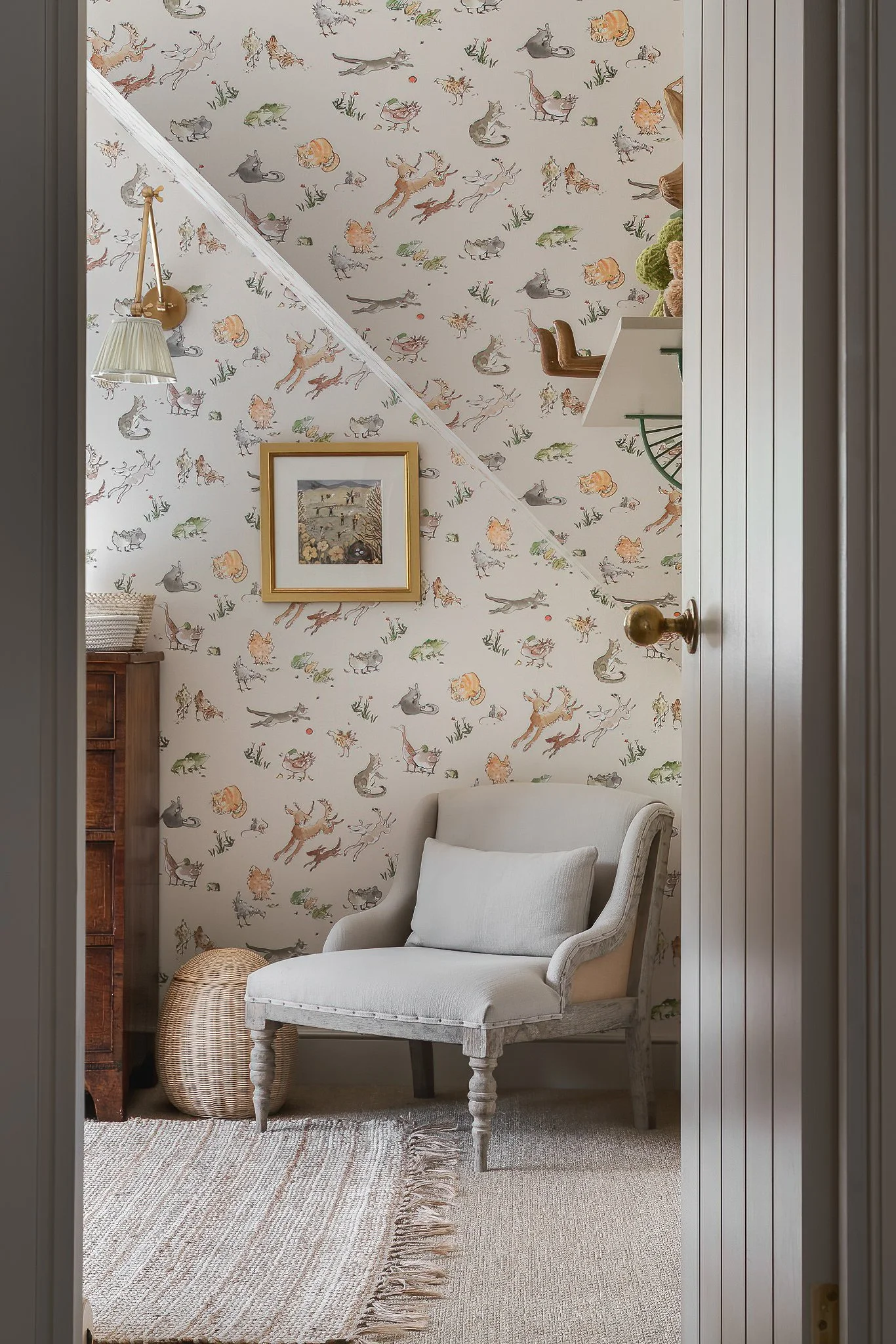 A cozy corner of a room with wallpaper featuring various animals, a white upholstered armchair with cushion, a woven basket on the floor, a small wooden dresser, and a framed picture on the wall.