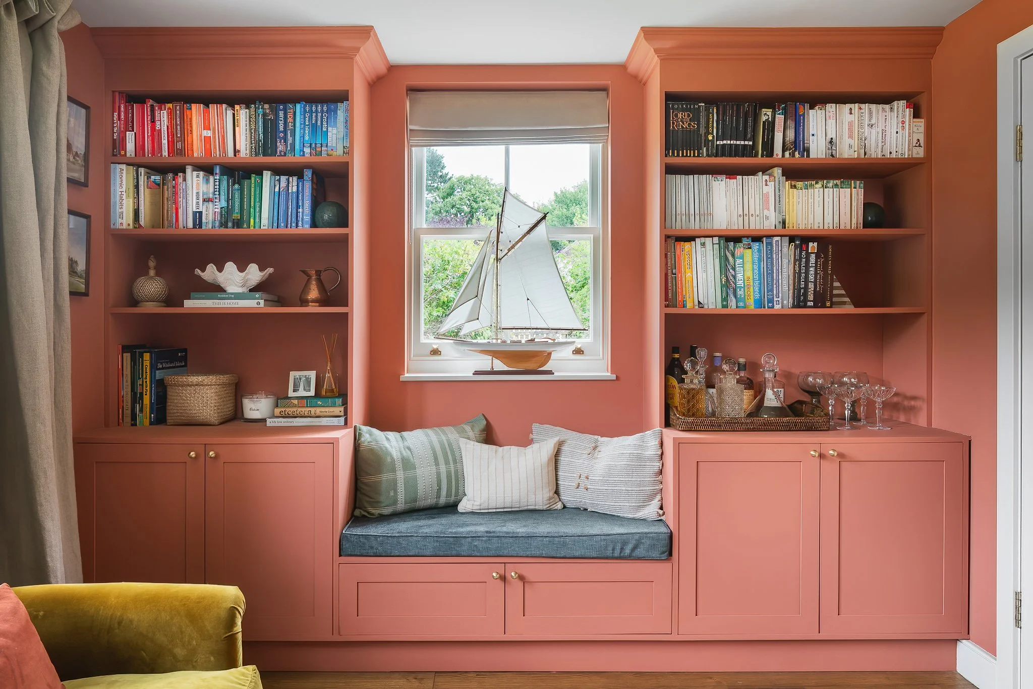 Bookcase with built-in window seat, pink cabinetry, and books, decorative objects, and glassware on the shelves, with a window and greenery outside.