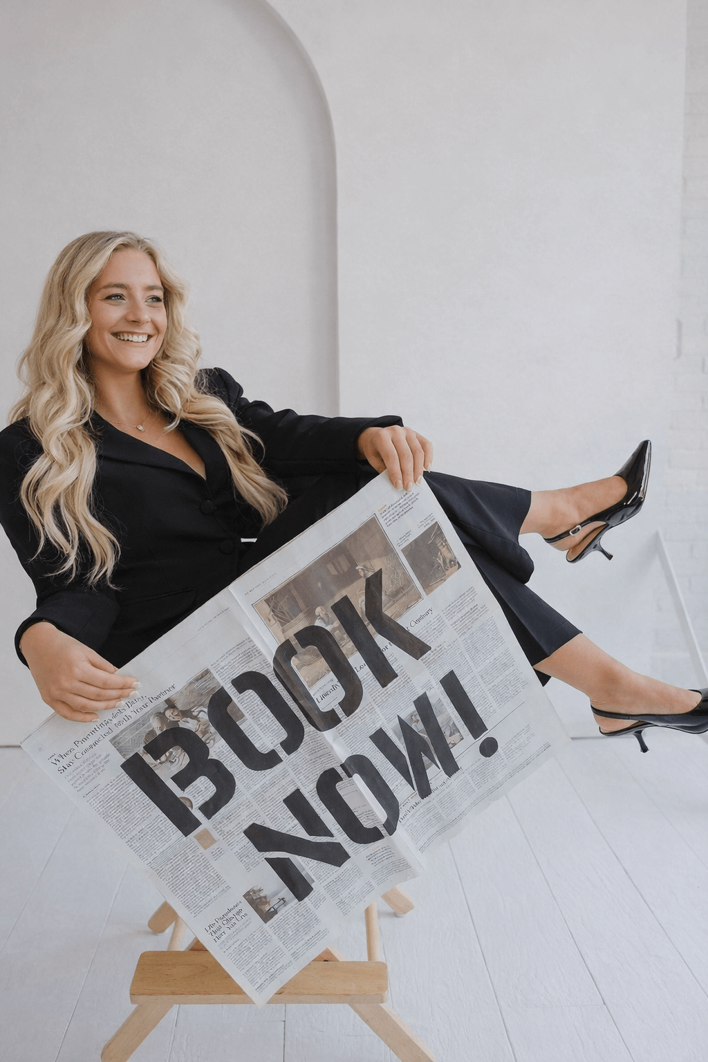 A woman in a black blazer and heels sitting on a wooden stool, smiling, while holding an upside-down newspaper with bold headlines.