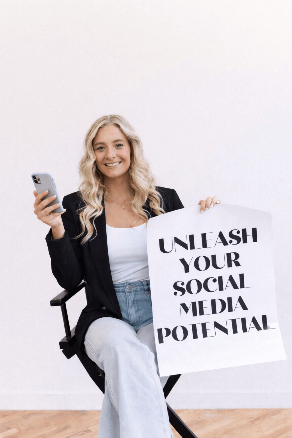 Young woman with blonde wavy hair smiling and holding a smartphone in one hand and a sign that says 'Unleash Your Social Media Potential'.